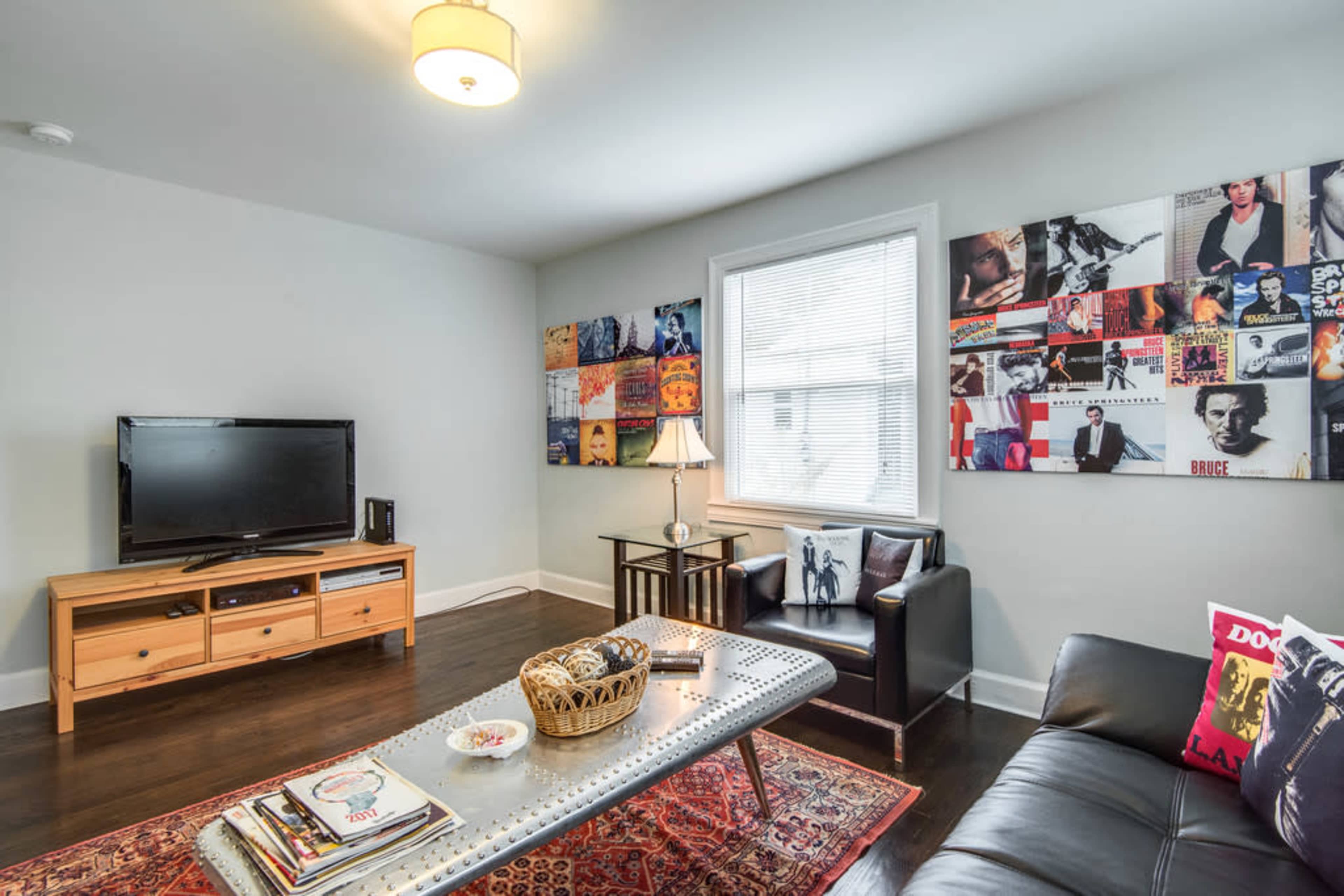 A living room with a television on a wooden stand, a coffee table with magazines, a black leather sofa, and a gallery wall featuring various album covers.