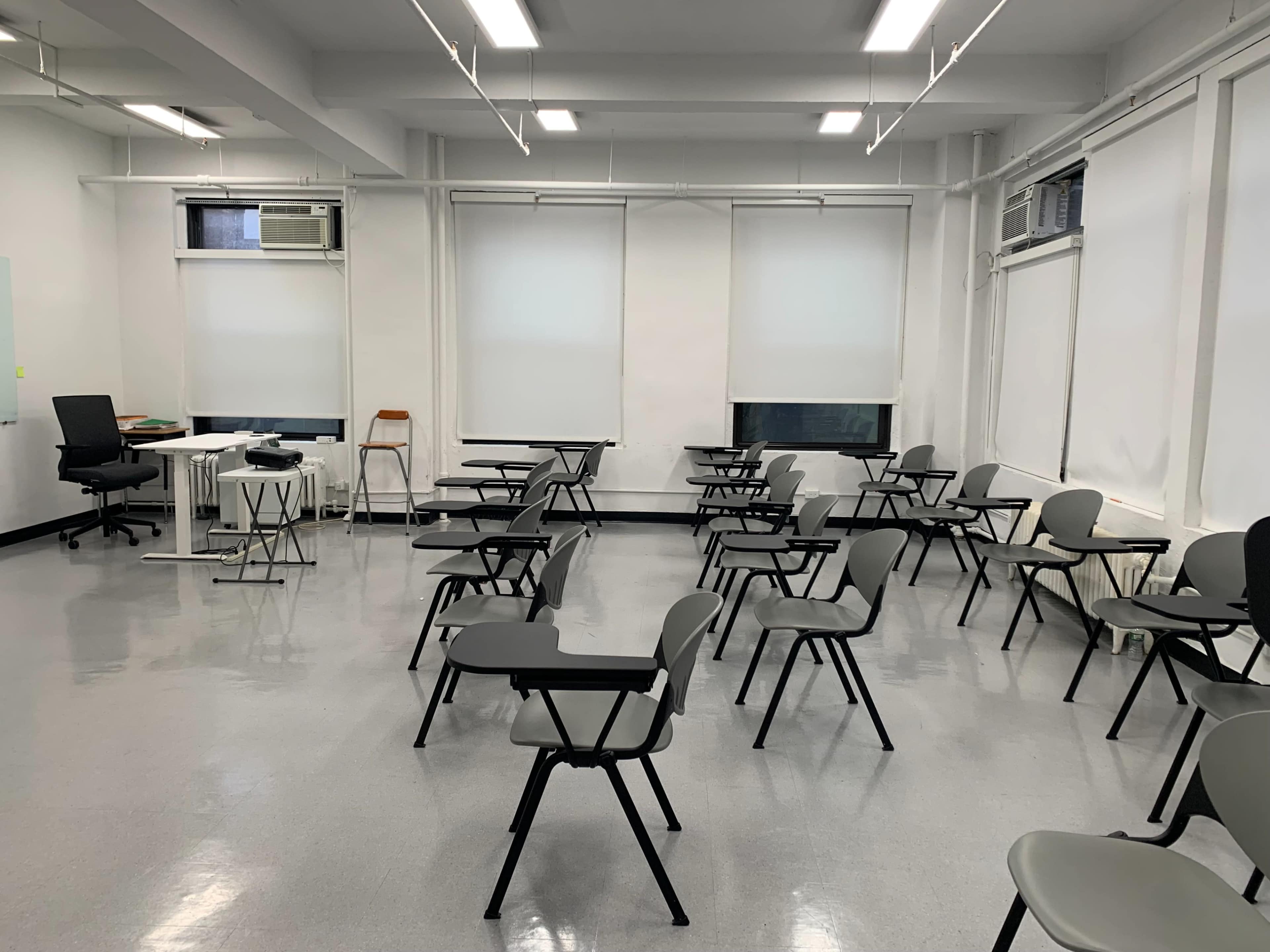 A classroom is arranged with individual desks and chairs spaced apart, and large windows with white blinds on two walls.