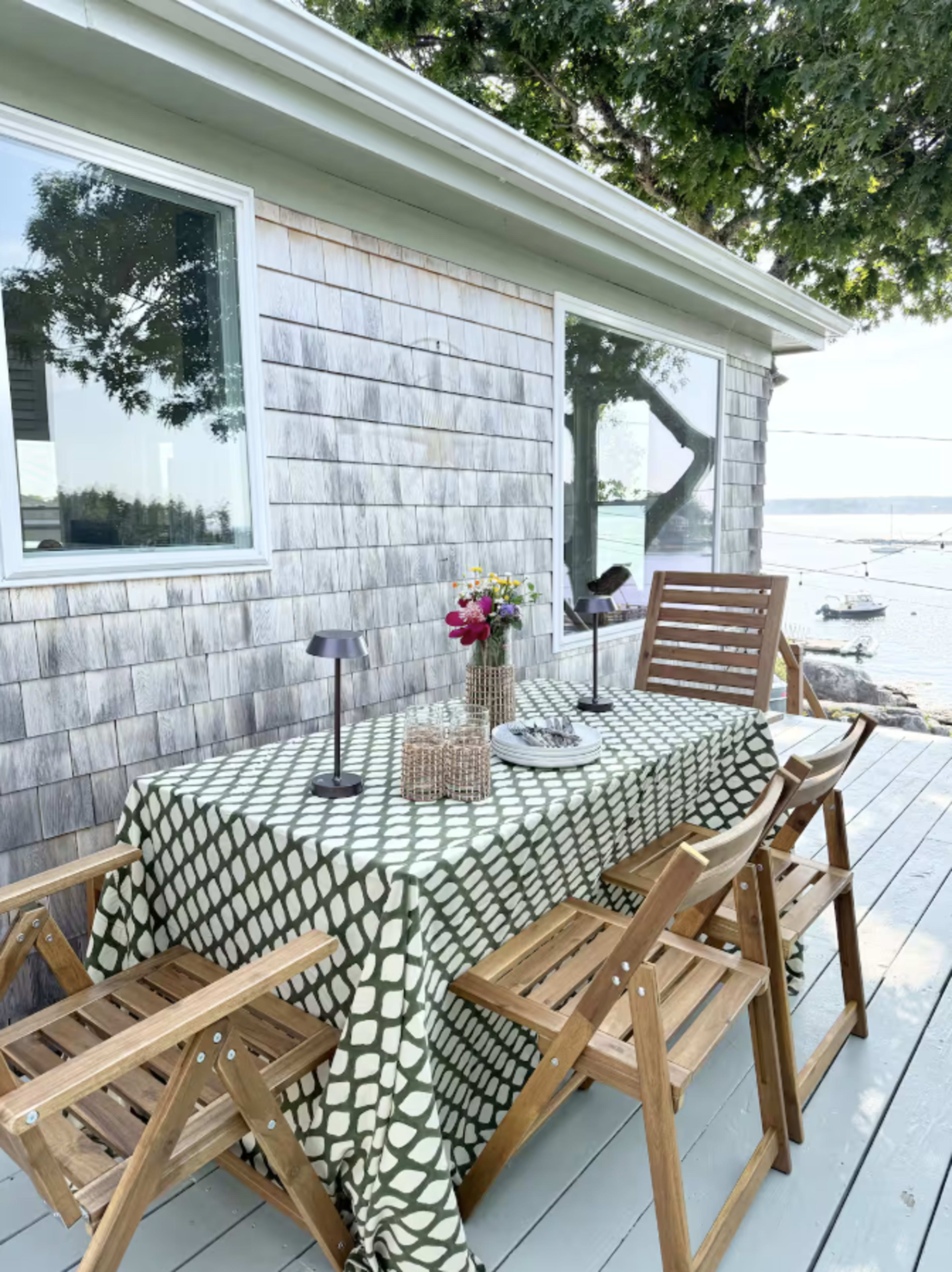 A wooden outdoor dining table set with a green and white patterned tablecloth and surrounded by folding chairs is positioned on a deck next to a house with shingled siding, overlooking a body of water.