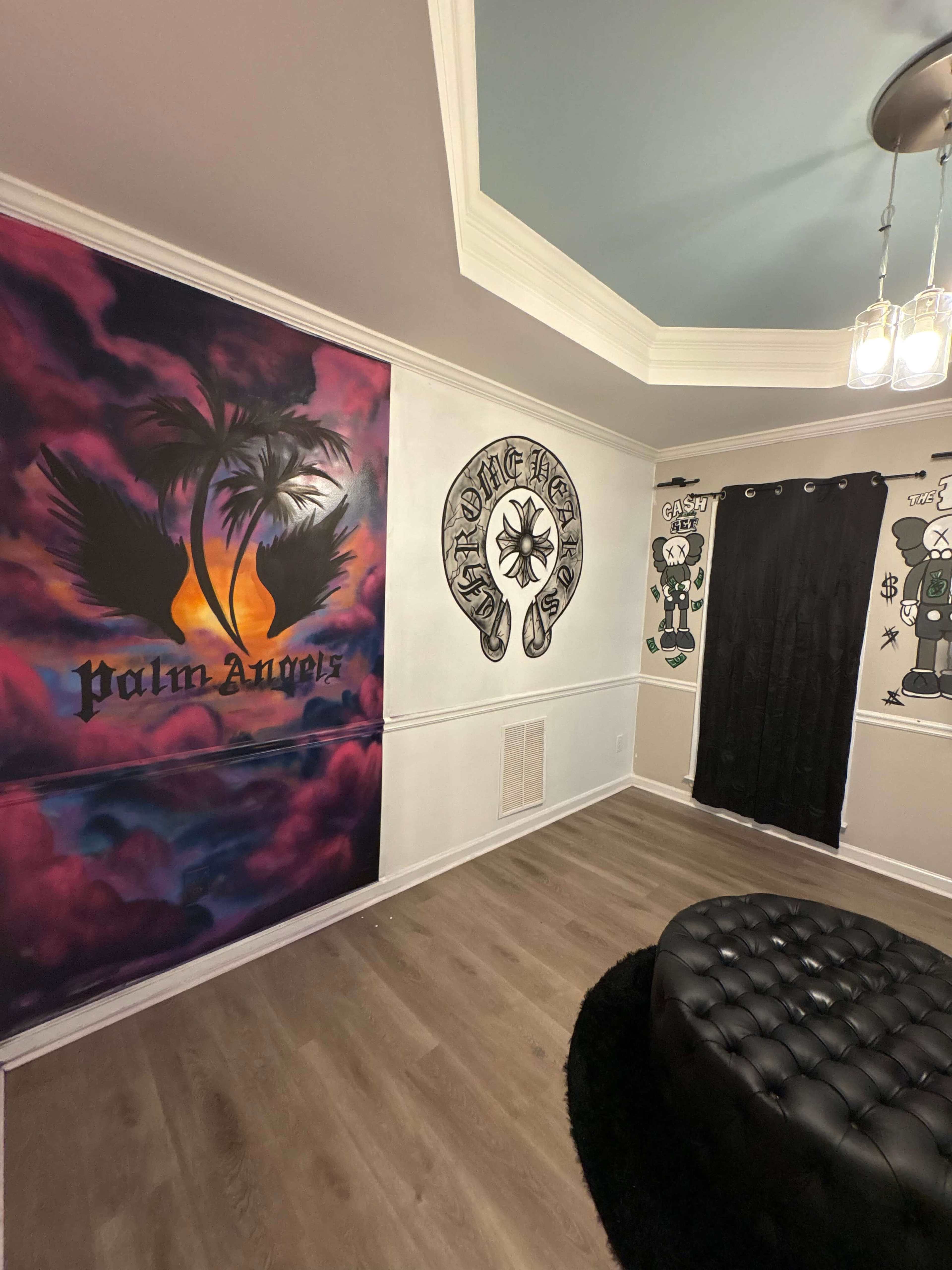 The image shows a room featuring a mural of palm trees and a sunset on one wall, intricate designs on another, a black curtain, and a tufted black ottoman on a wooden floor.