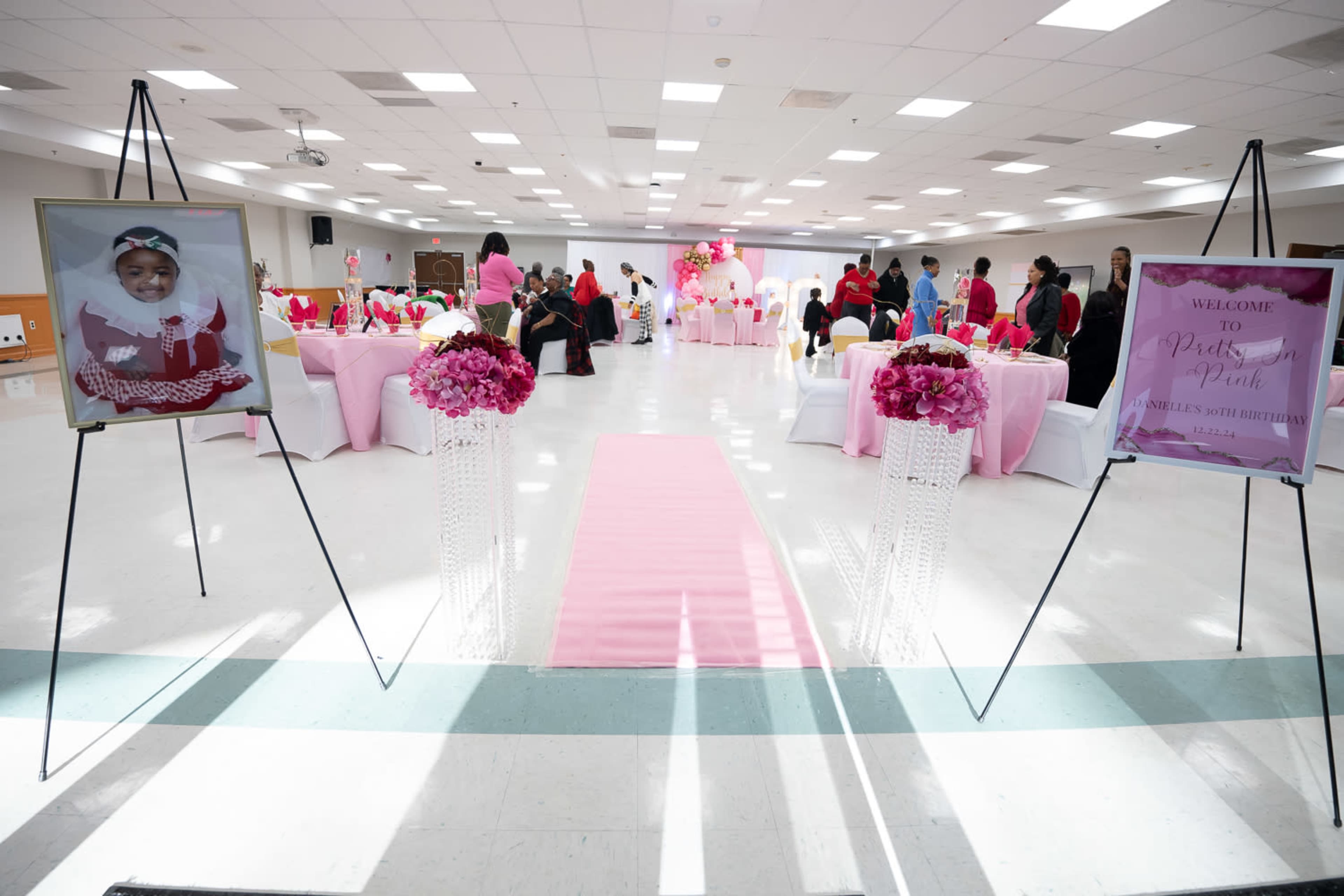 The image shows a decorated event space for a birthday celebration, featuring pink table settings, floral arrangements, and signs welcoming guests.