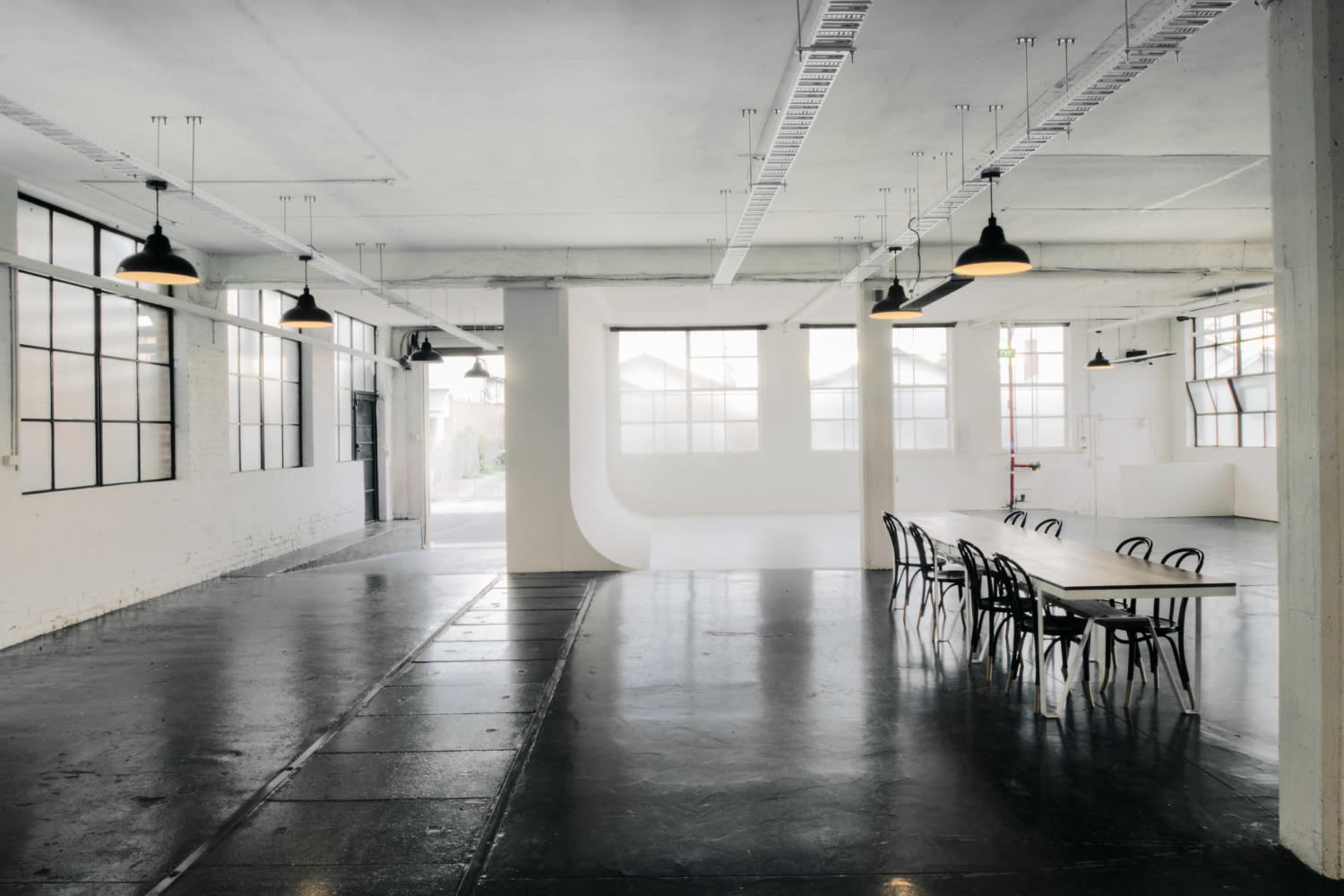 The image shows a spacious, industrial-style room with large windows, high ceilings, and a long table surrounded by black chairs.