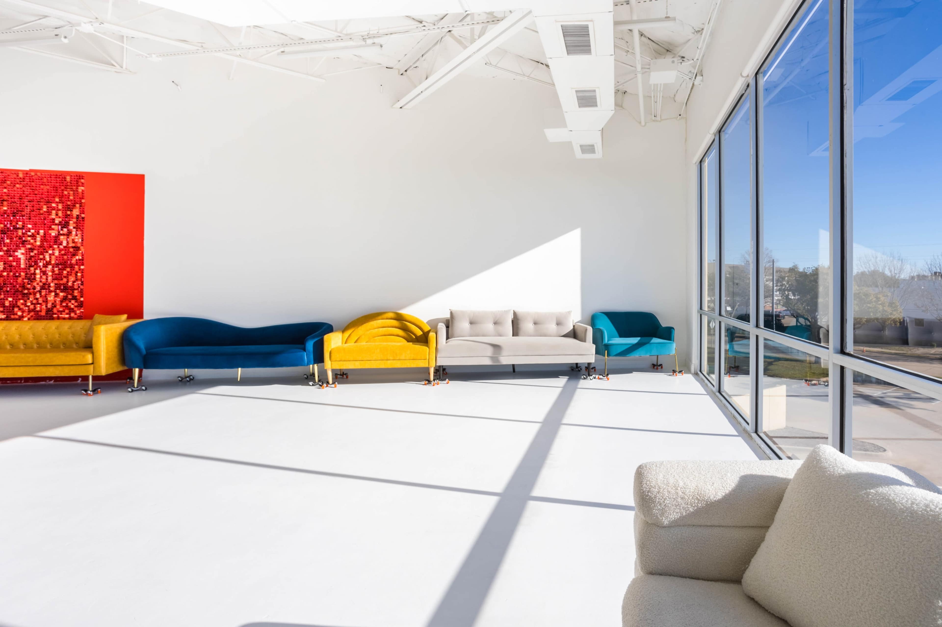 The image shows a spacious, well-lit room with colorful couches arranged along one wall and large windows allowing natural light to enter.