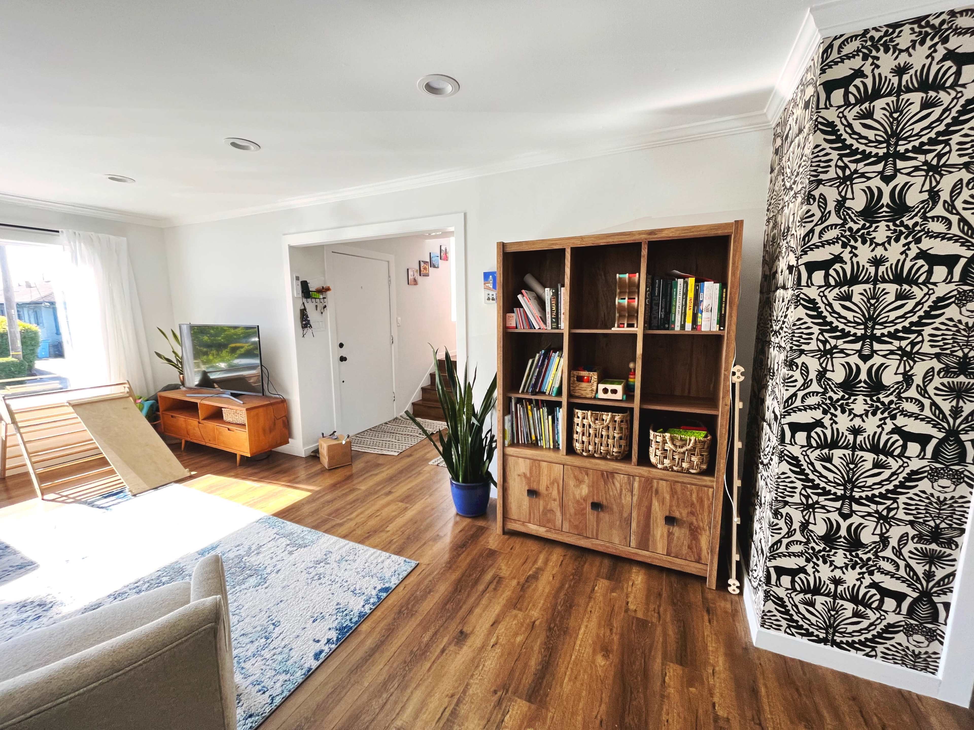 The living room features a large bookshelf, a television stand, and a door leading outside, with a patterned wallpaper accent wall.