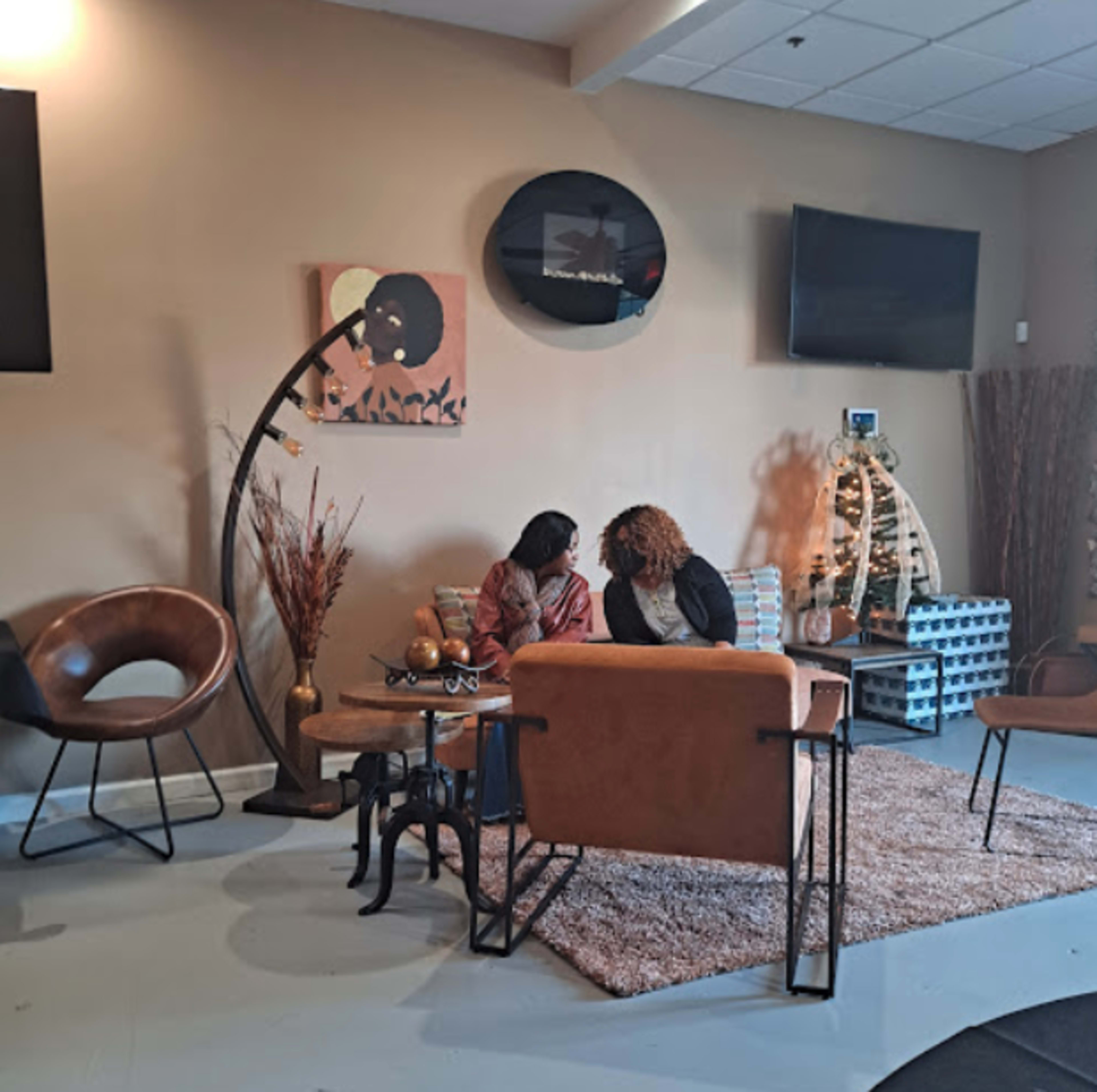 Two women sit on a couch in a modern lounge area decorated with plants, artwork, and television screens.