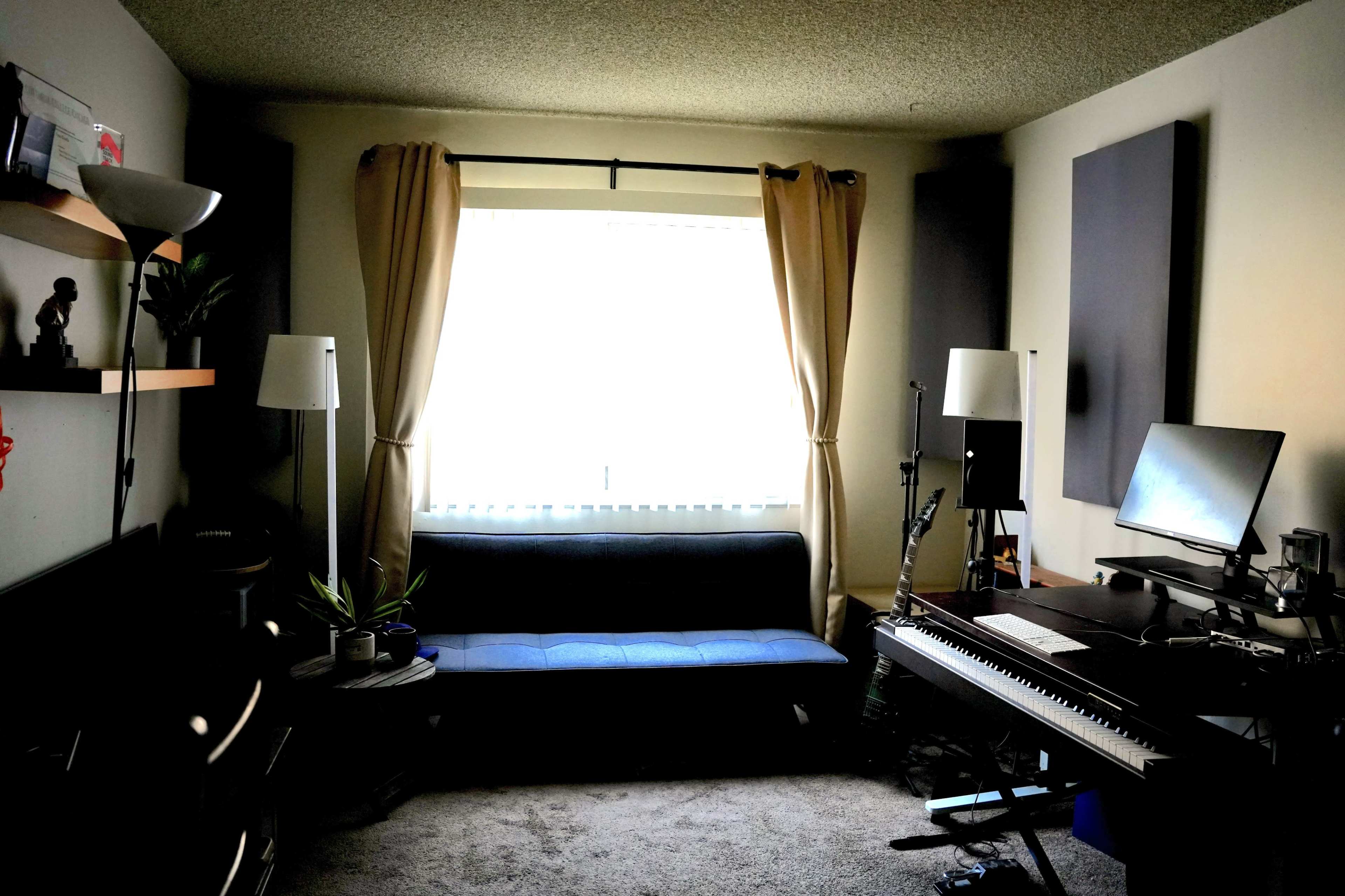 The image depicts a cozy room featuring a blue couch, a computer desk with a monitor, and two floor lamps, illuminated by natural light coming through a window with beige curtains.