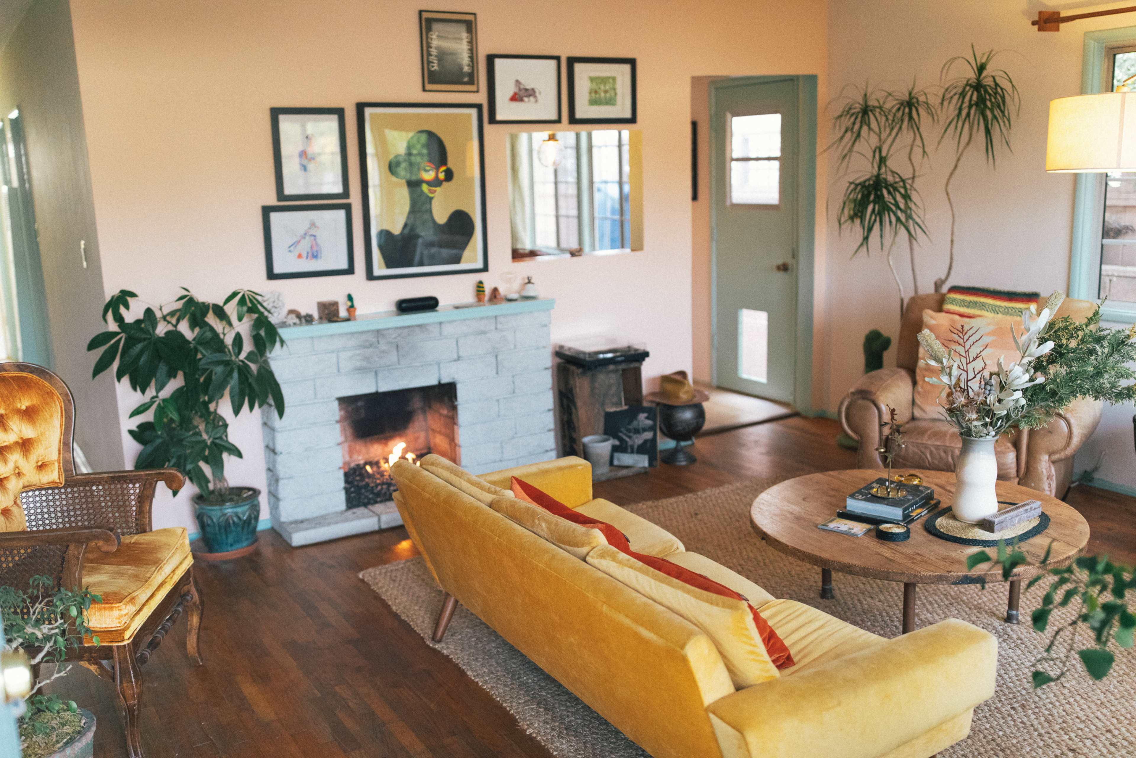 The scene features a cozy living room with a yellow couch, a wooden coffee table, an art-adorned wall, a fireplace, and various plants.