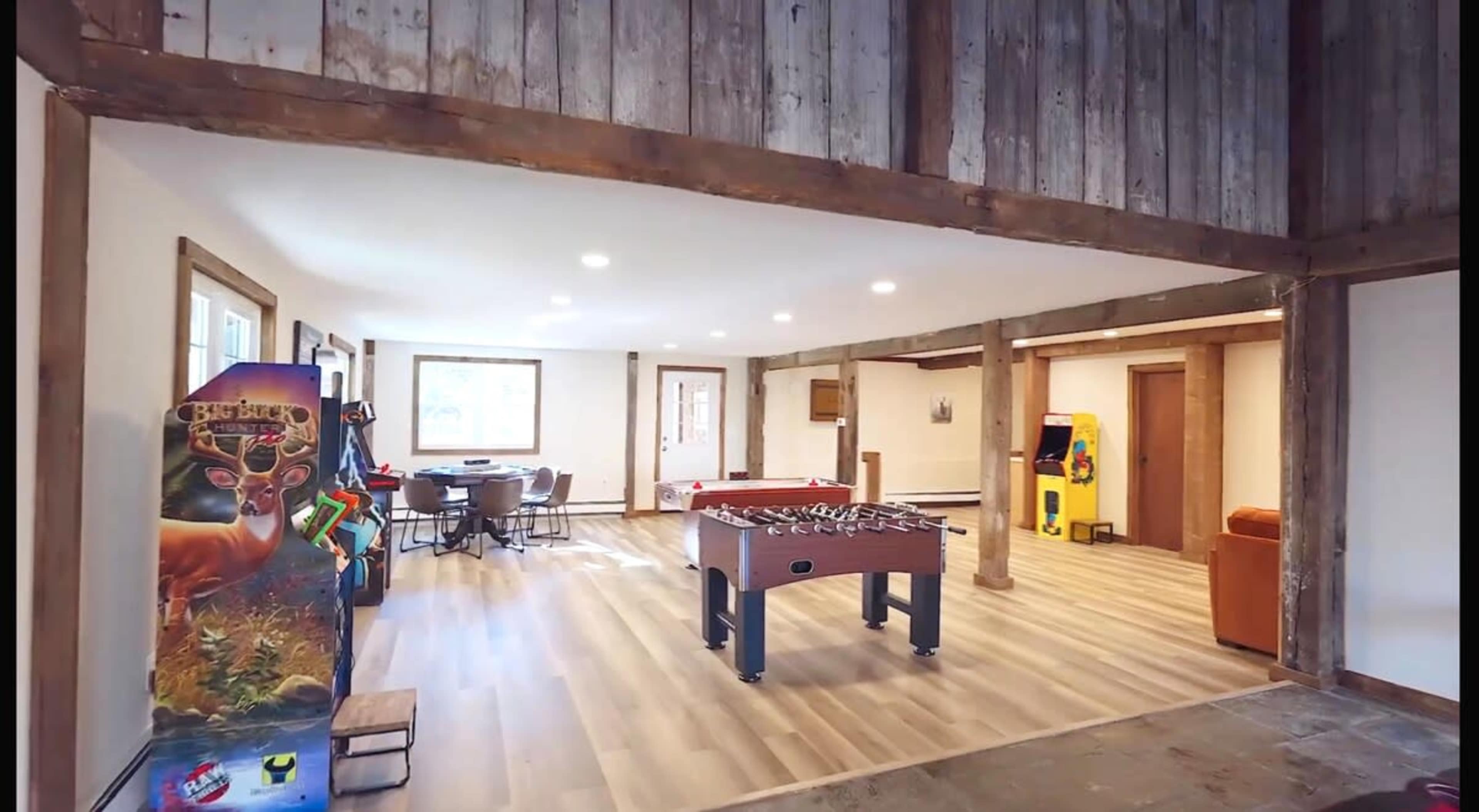 The image shows a spacious game room featuring a foosball table, arcade machines, and a dining area with a round table surrounded by chairs.