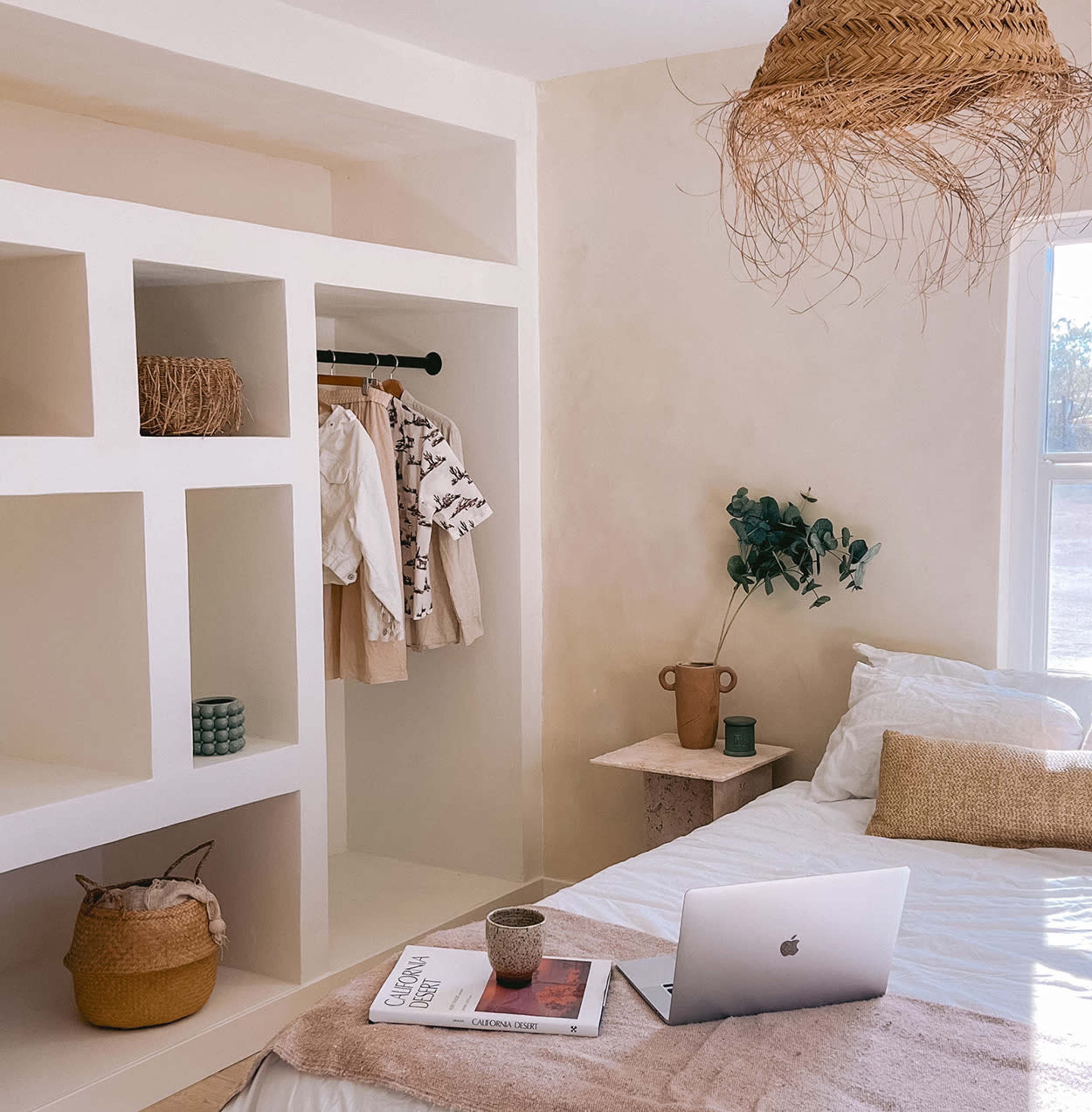 A neatly arranged bedroom features a bed with a laptop on it, a clothing rack with hanging garments, and shelves displaying decorative items.