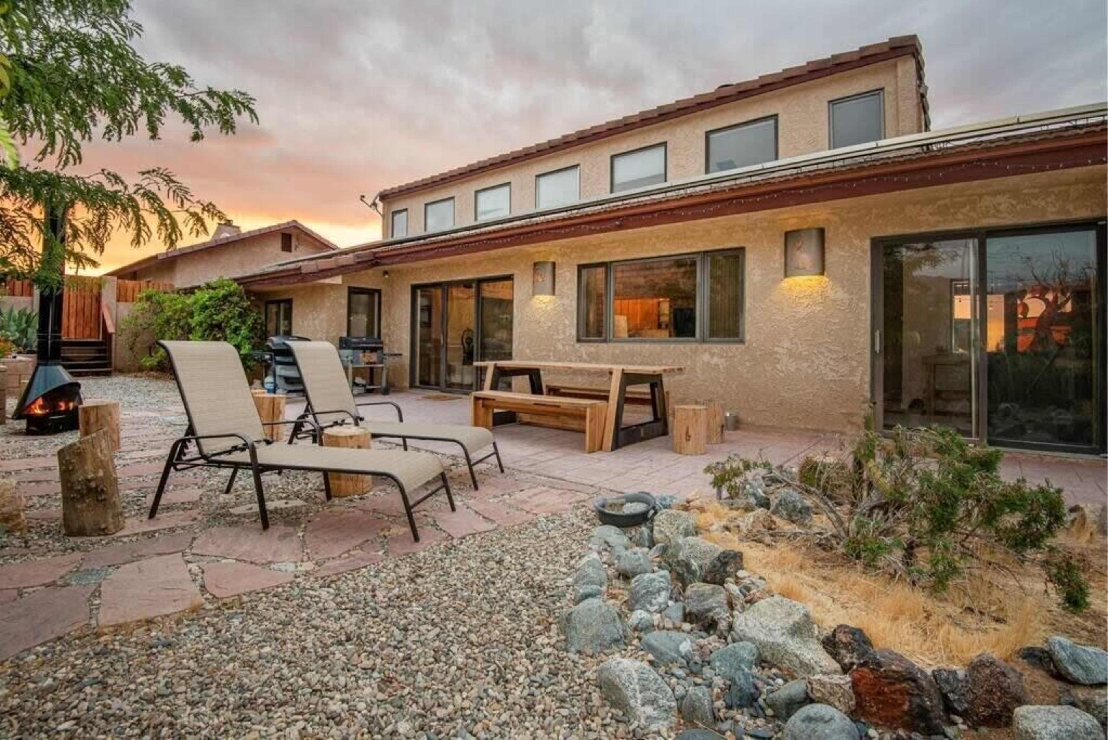 A desert-style backyard features lounge chairs, a wooden dining table, and a stone pathway leading to a two-story house with large windows.