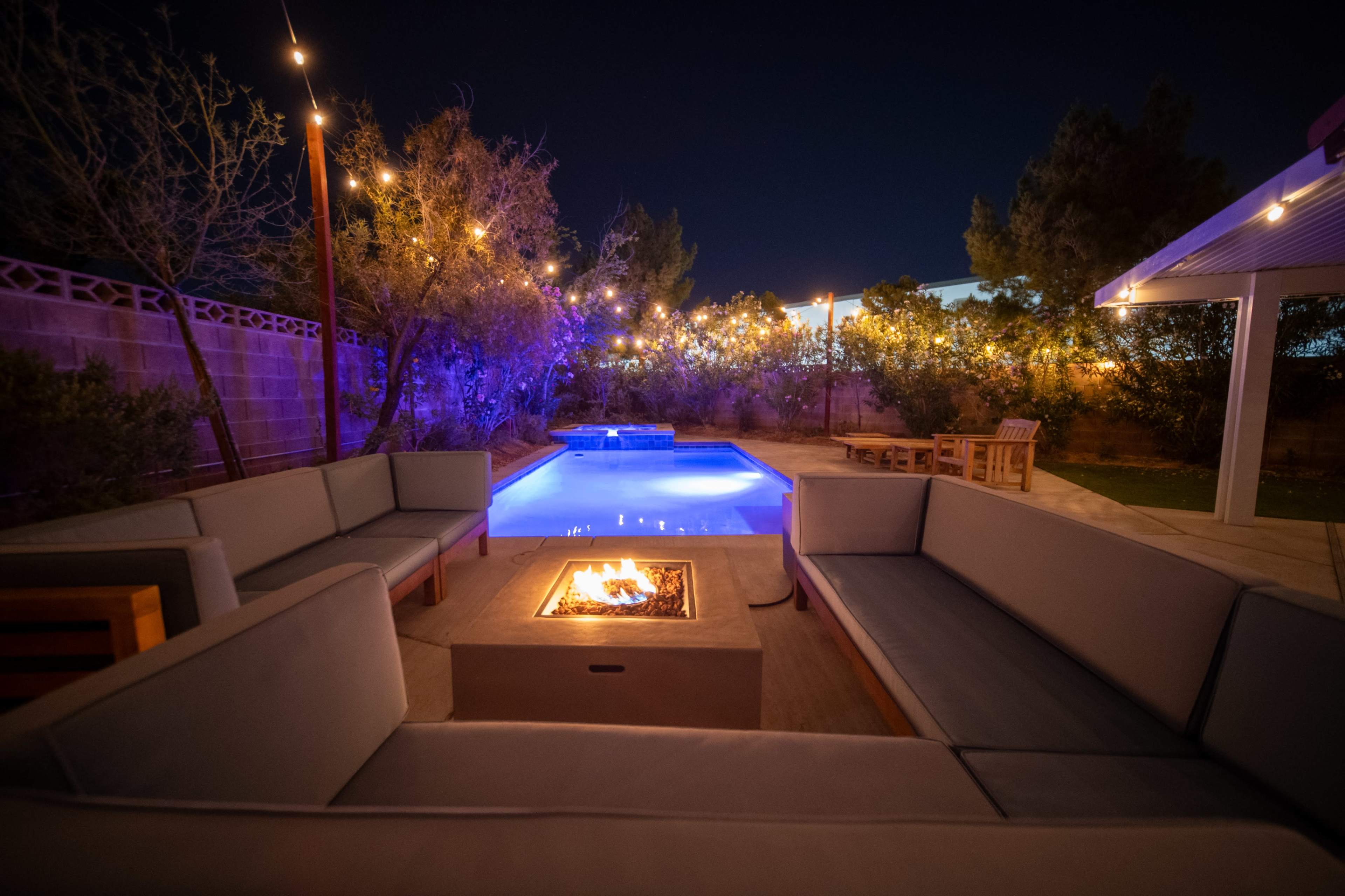 A cozy outdoor seating area surrounds a fire pit next to a illuminated swimming pool, under a night sky adorned with string lights.
