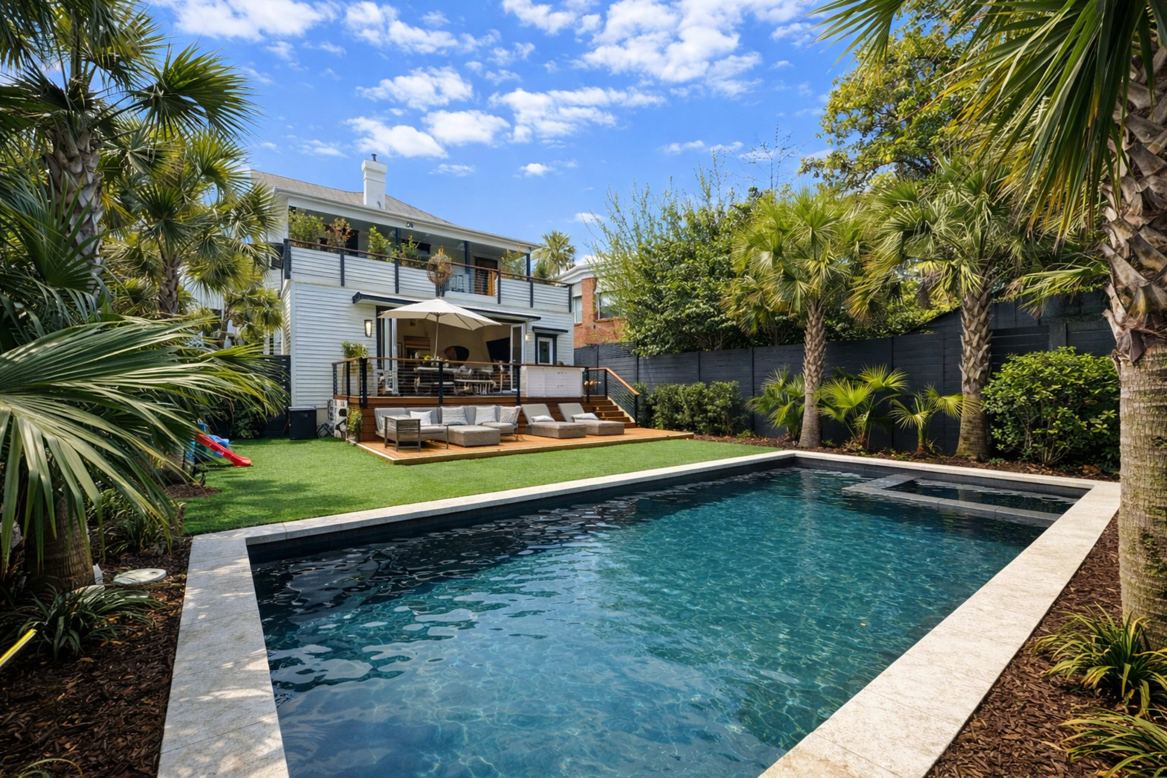 The image shows a backyard with a rectangular swimming pool surrounded by palm trees, a grassy area, and a wooden deck with lounge furniture and an umbrella.