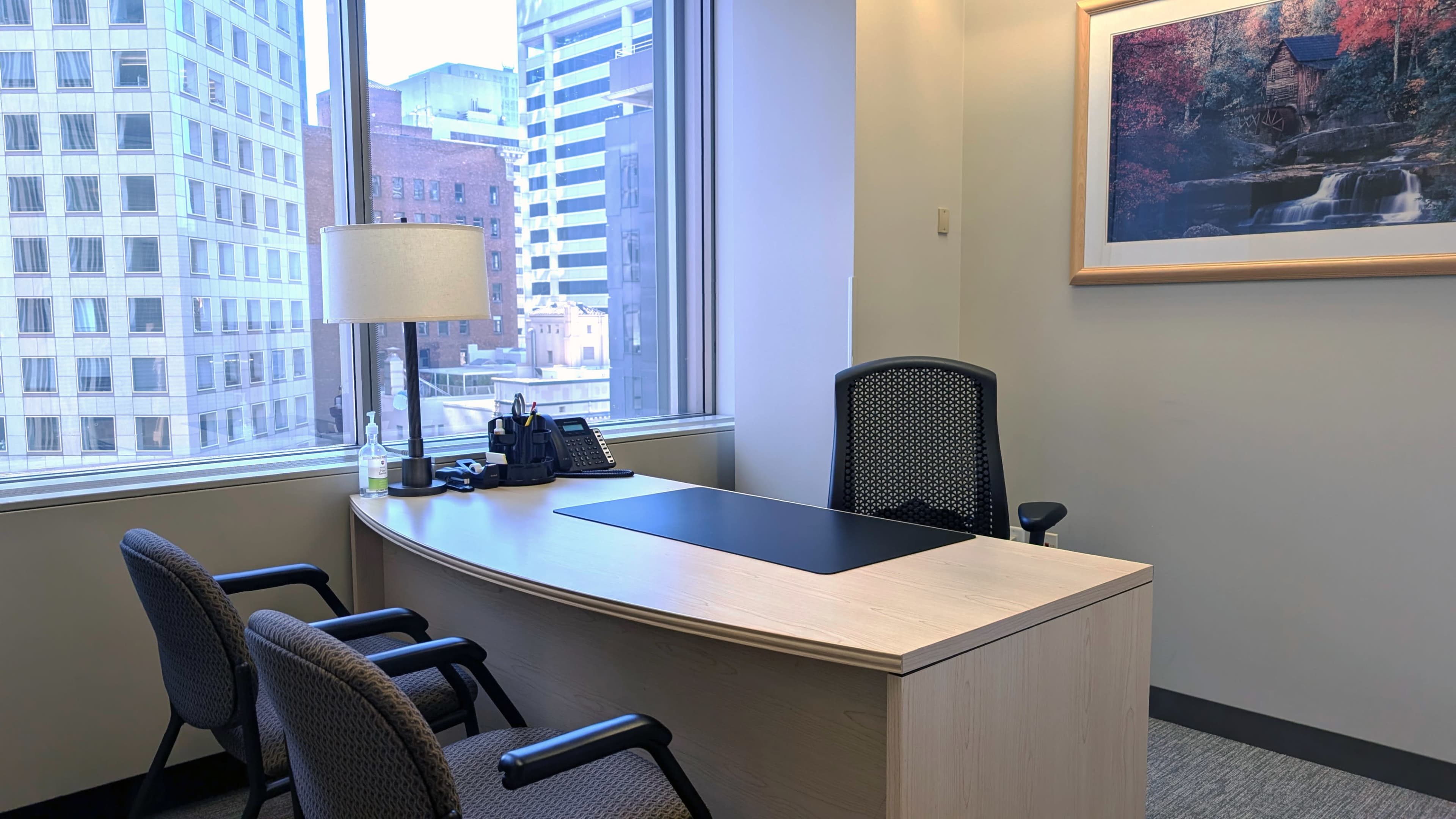 The image shows an office space featuring a curved wooden desk, a chair behind it, and two chairs in front, with large windows overlooking city buildings.