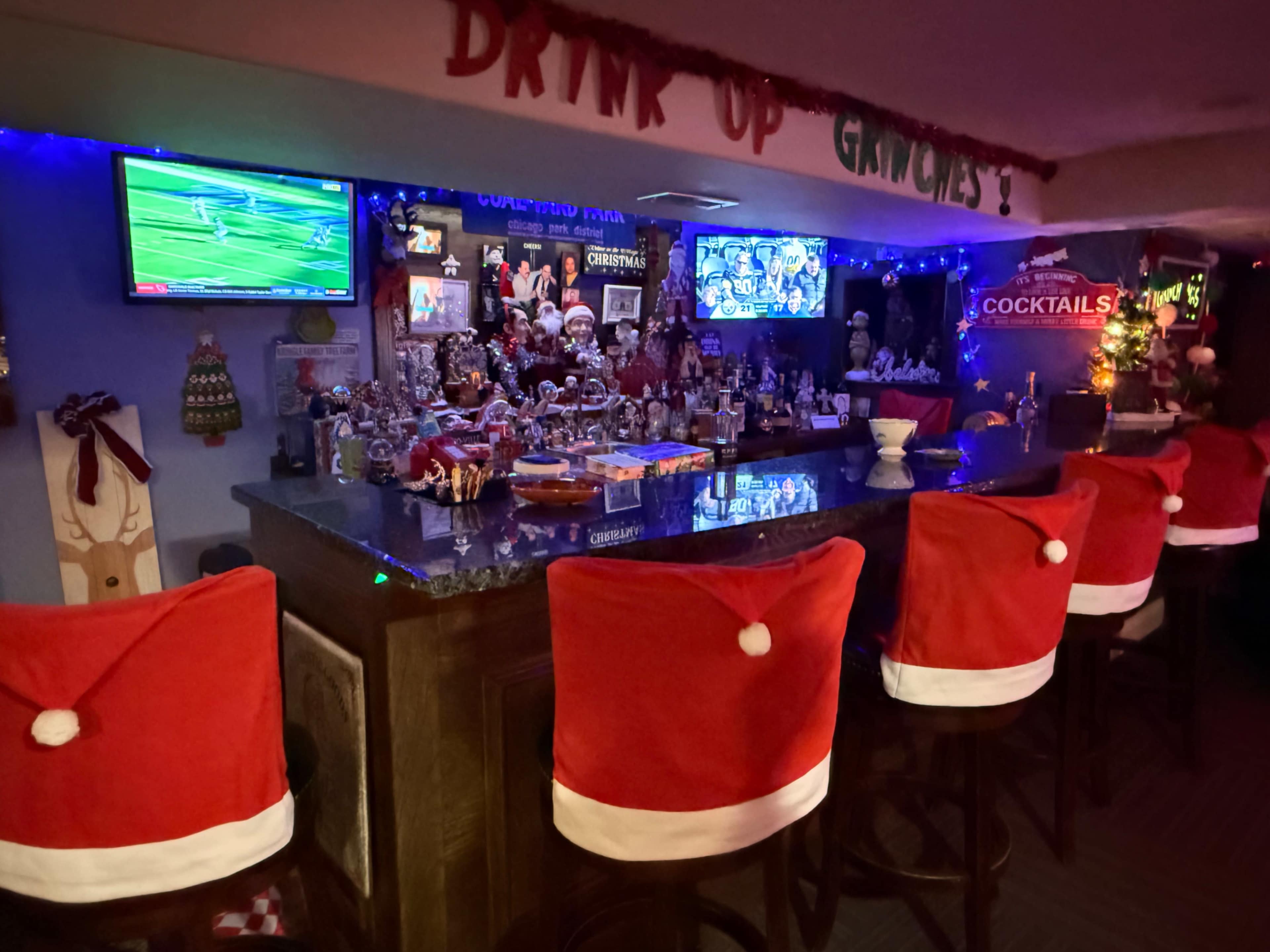 A festive bar area features a counter with eight stools adorned in Santa hat covers, surrounded by holiday decorations and multiple screens displaying sports.