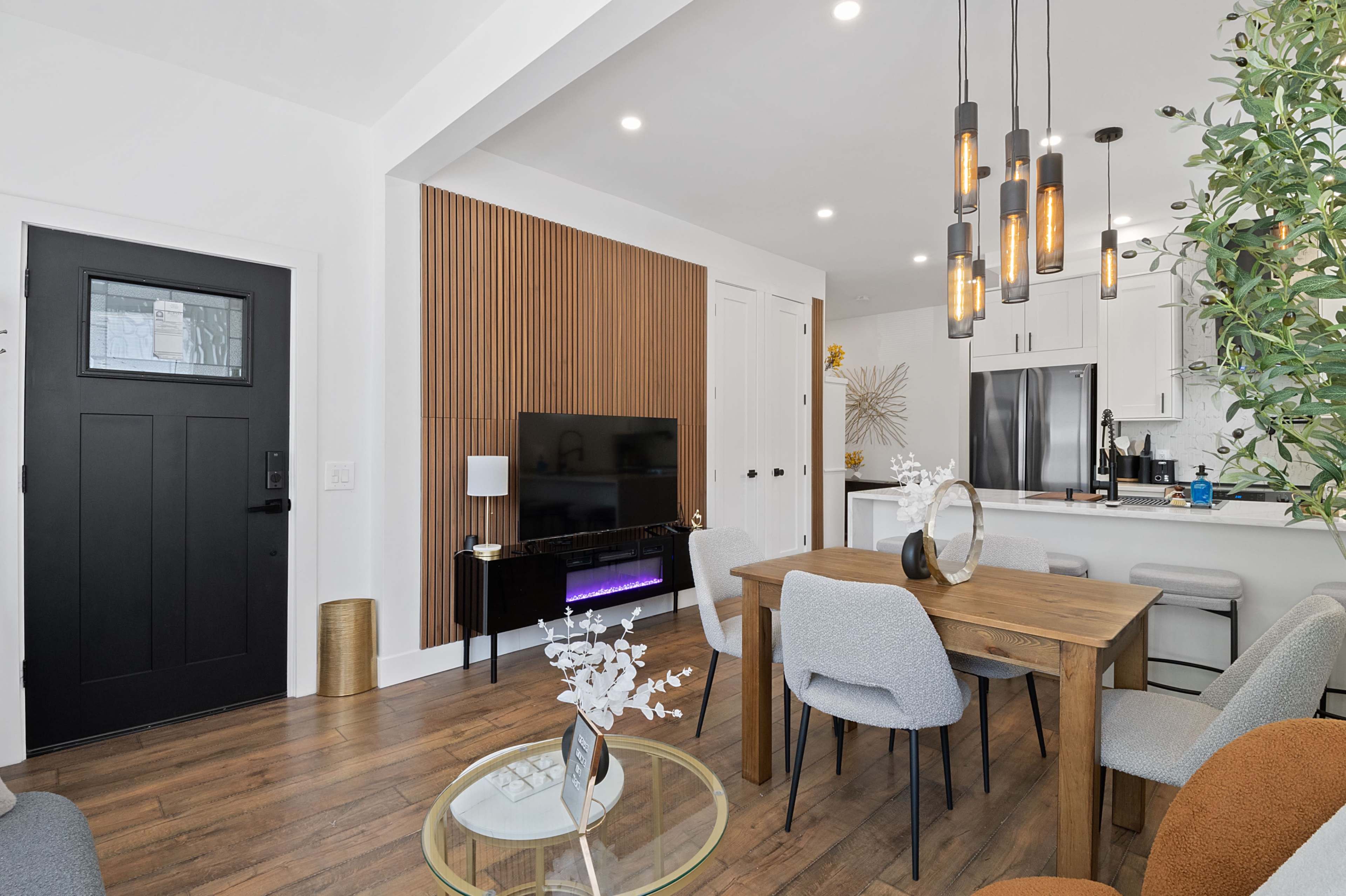 A modern living area features a wooden dining table surrounded by chairs, a television mounted on a textured wall, and a kitchen visible in the background.