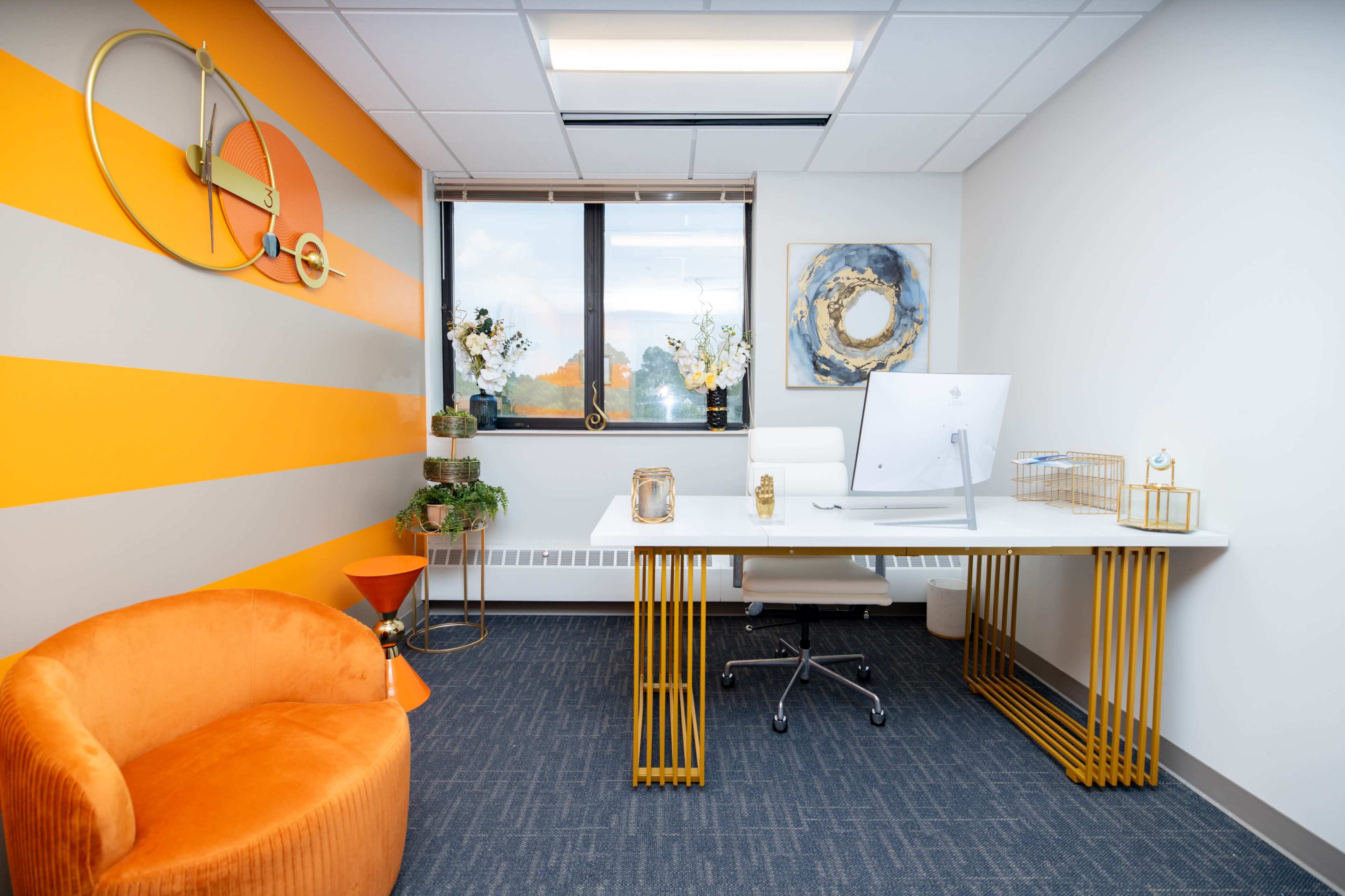 A modern office space featuring a white desk with a computer, an orange accent wall, a plush orange chair, and decorative plants near the window.