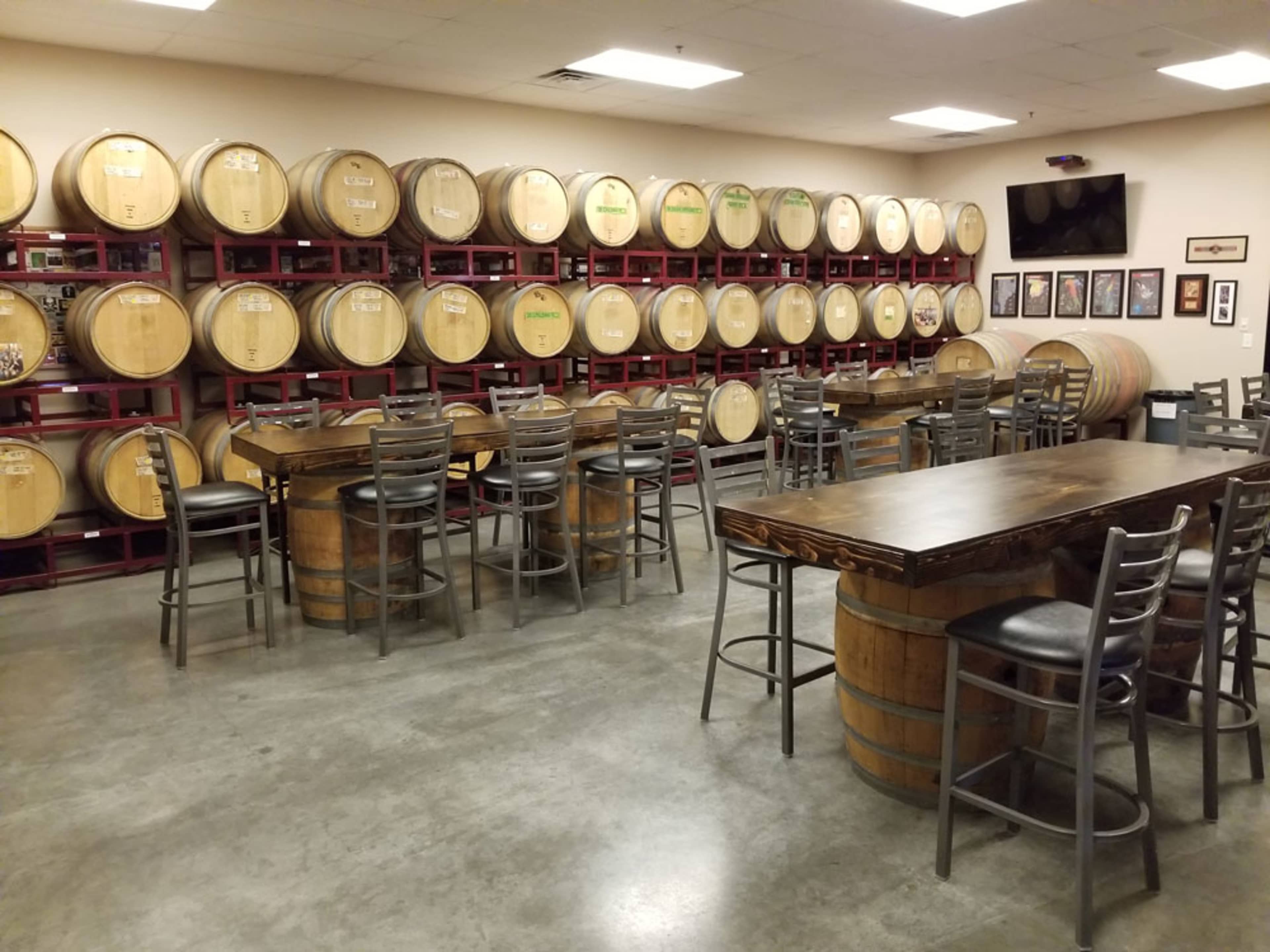 A wine tasting room with wooden barrels along the wall and tables with high stools arranged in the center.