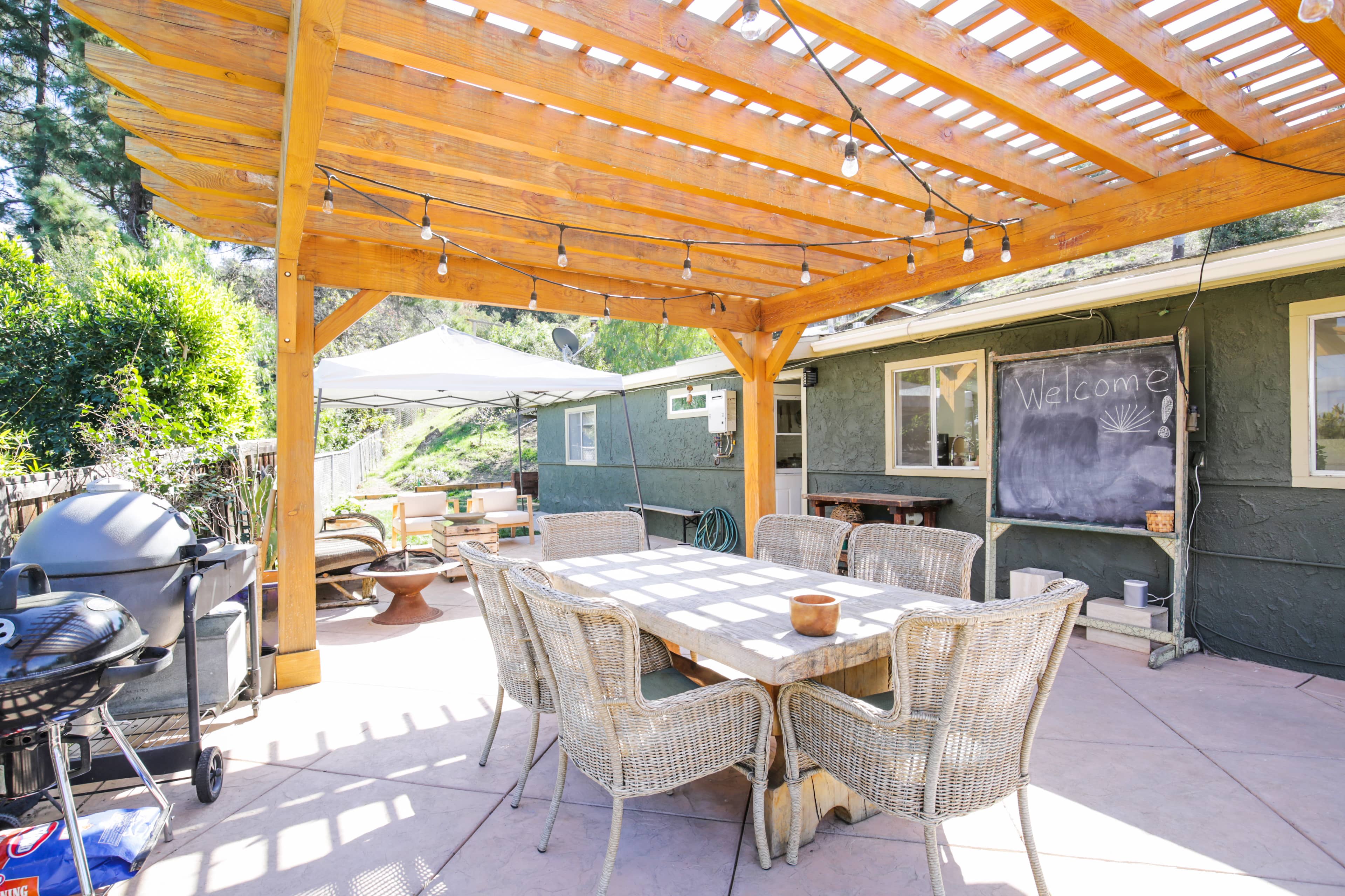 A patio area with a wooden pergola, dining table, and a grill, adjacent to a house surrounded by greenery.