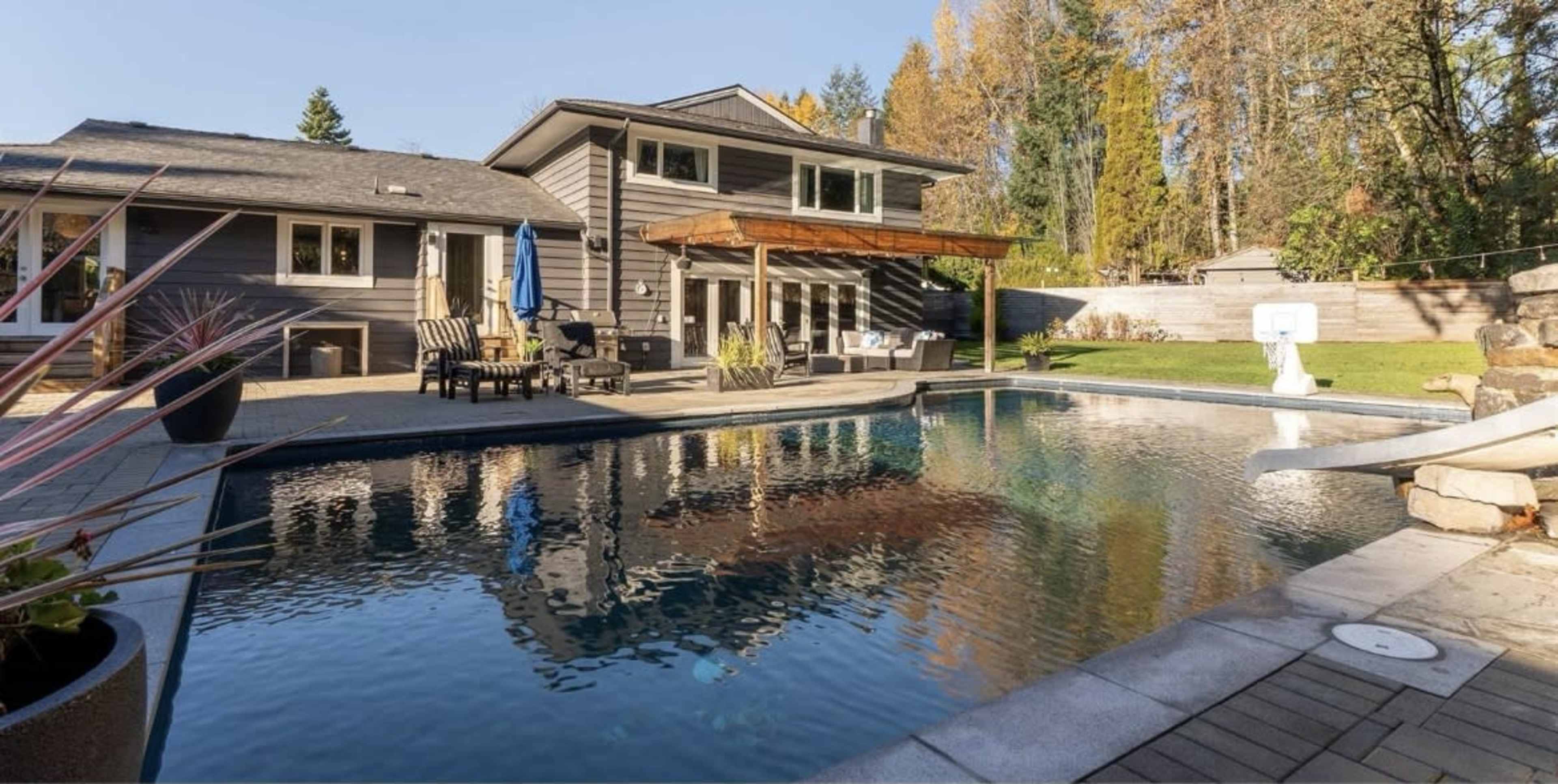 The image shows a modern home with a swimming pool surrounded by a wooden deck, patio furniture, and landscaped gardens.