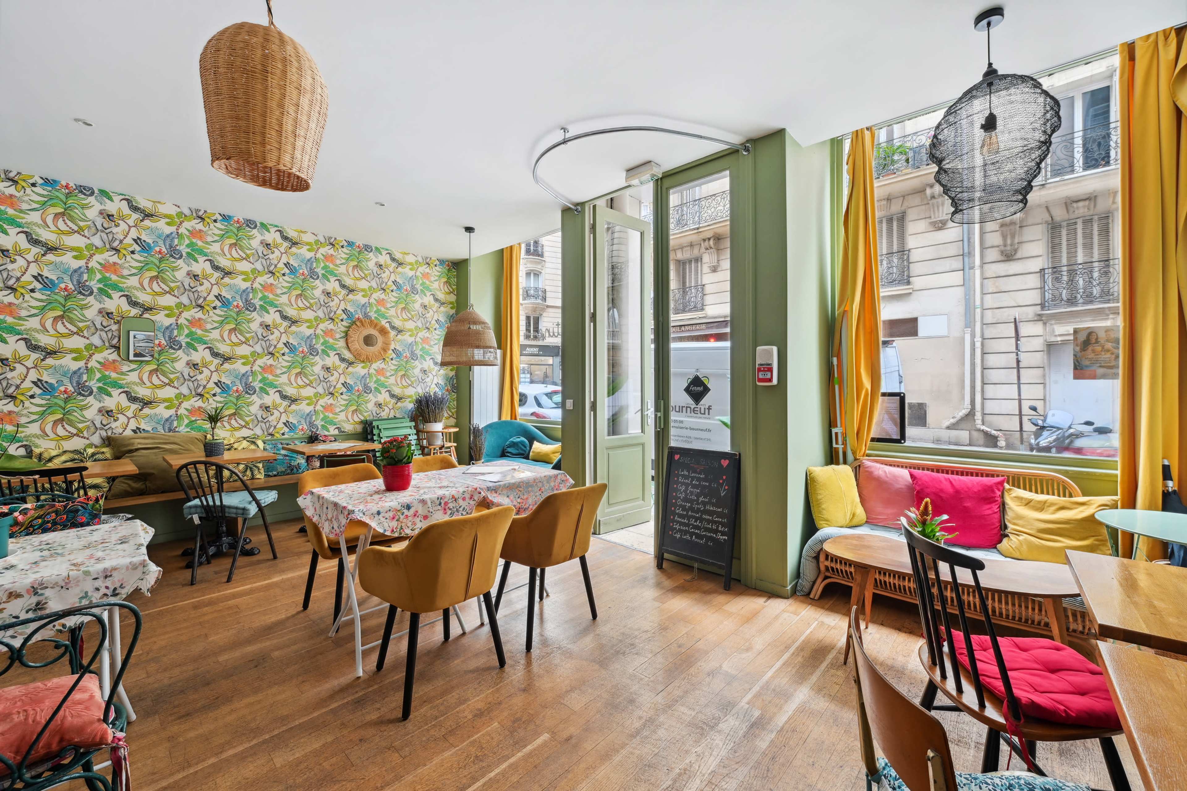 The image depicts a cozy café interior featuring colorful floral wallpaper, wooden flooring, various seating arrangements, and large windows that allow natural light to stream in.
