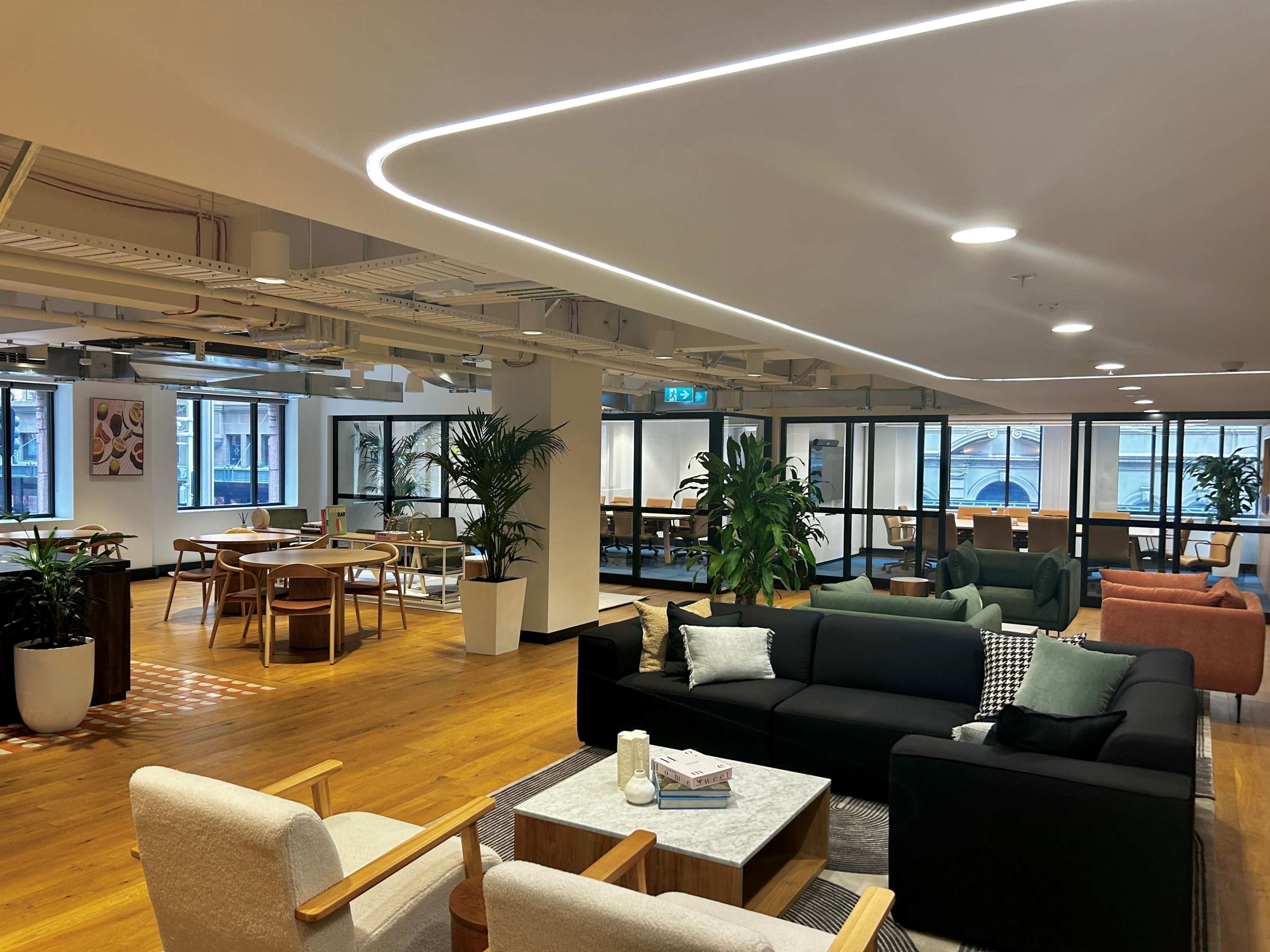 A modern office space features a lounge area with sofas, a coffee table, and plants, alongside dining tables and large windows.