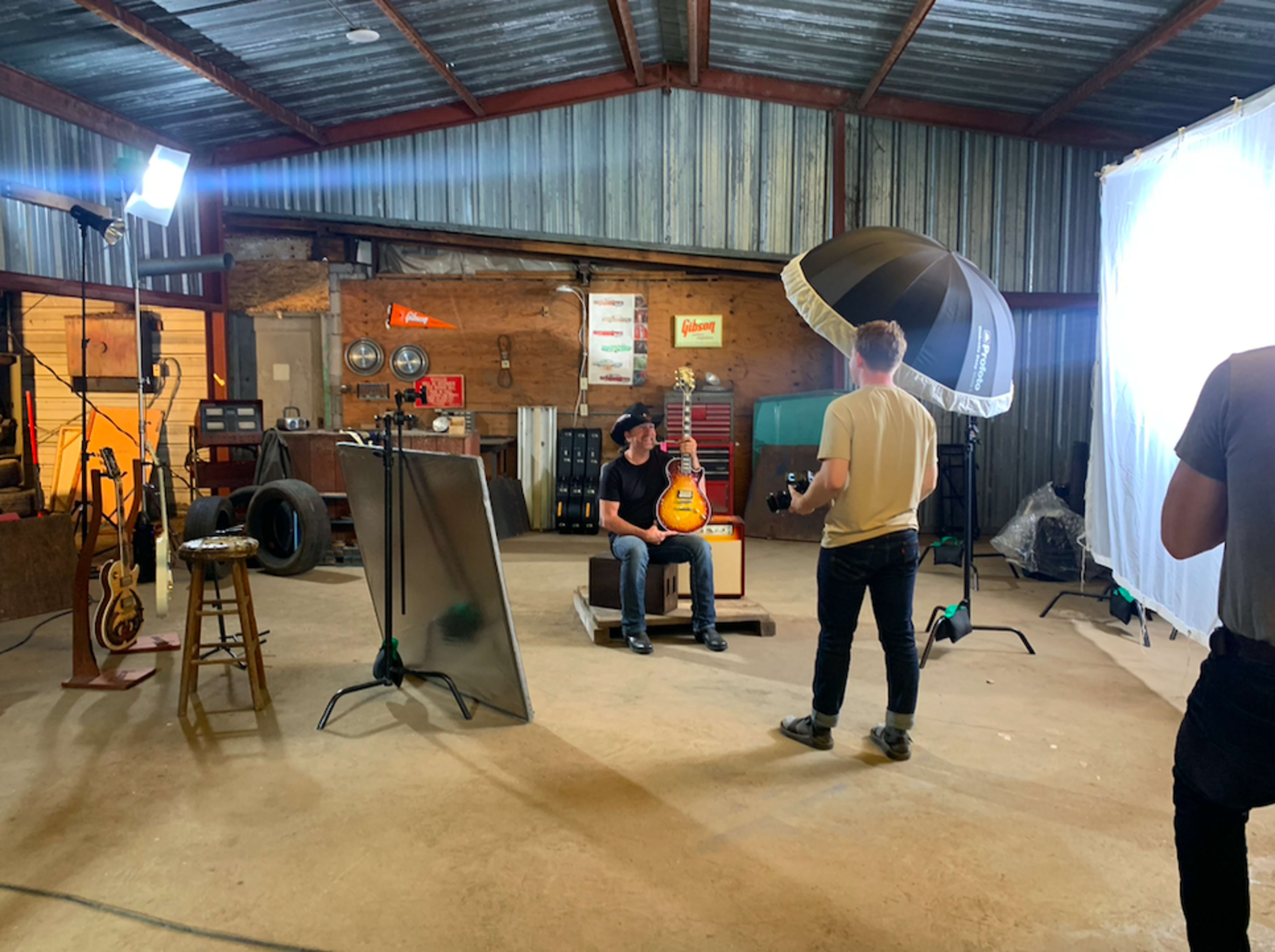 A person sits on a stool with an electric guitar in a dimly lit workshop, while another person adjusts lighting equipment nearby.