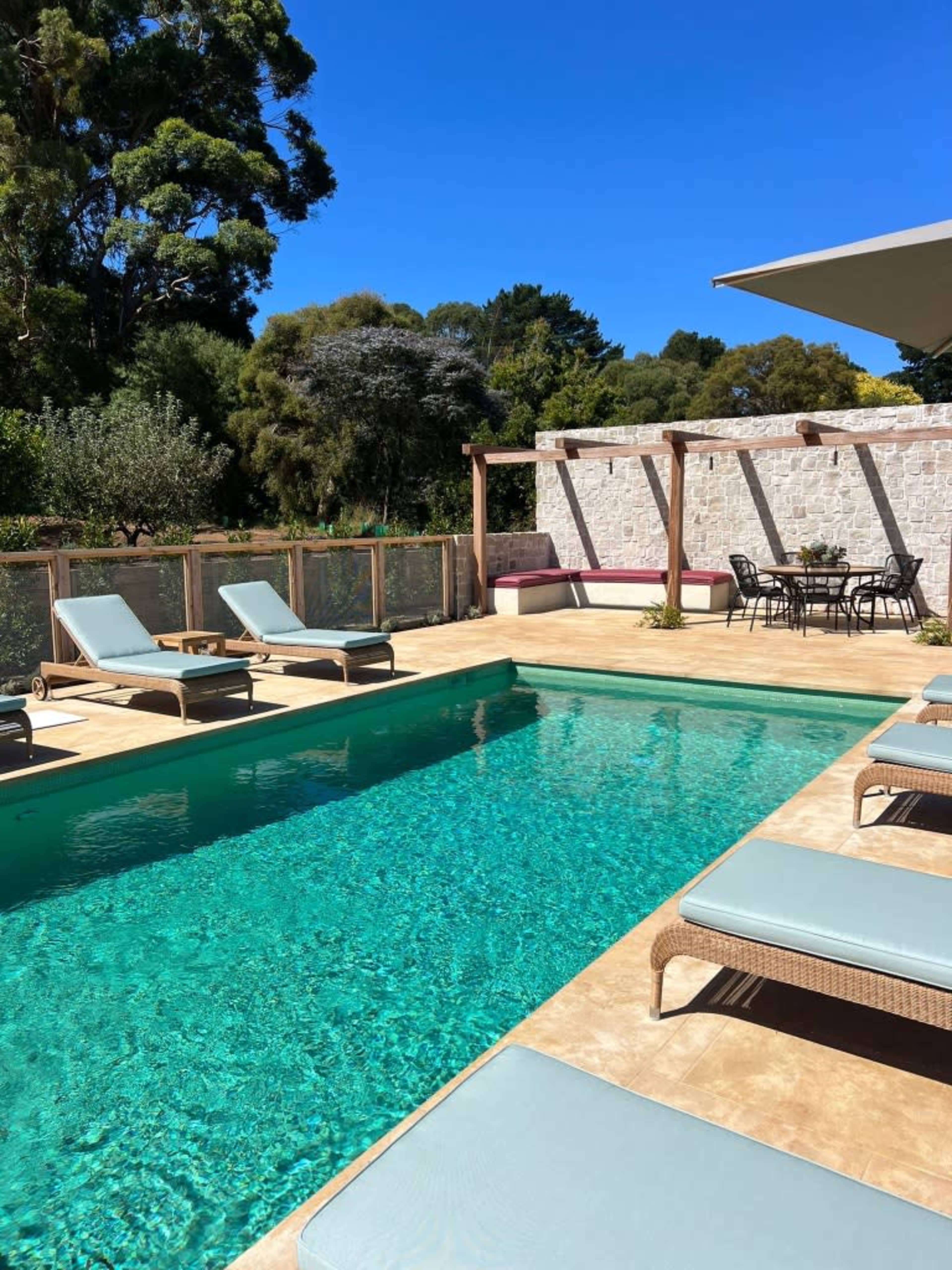 A swimming pool surrounded by lounge chairs, with a dining area and landscaping in the background.