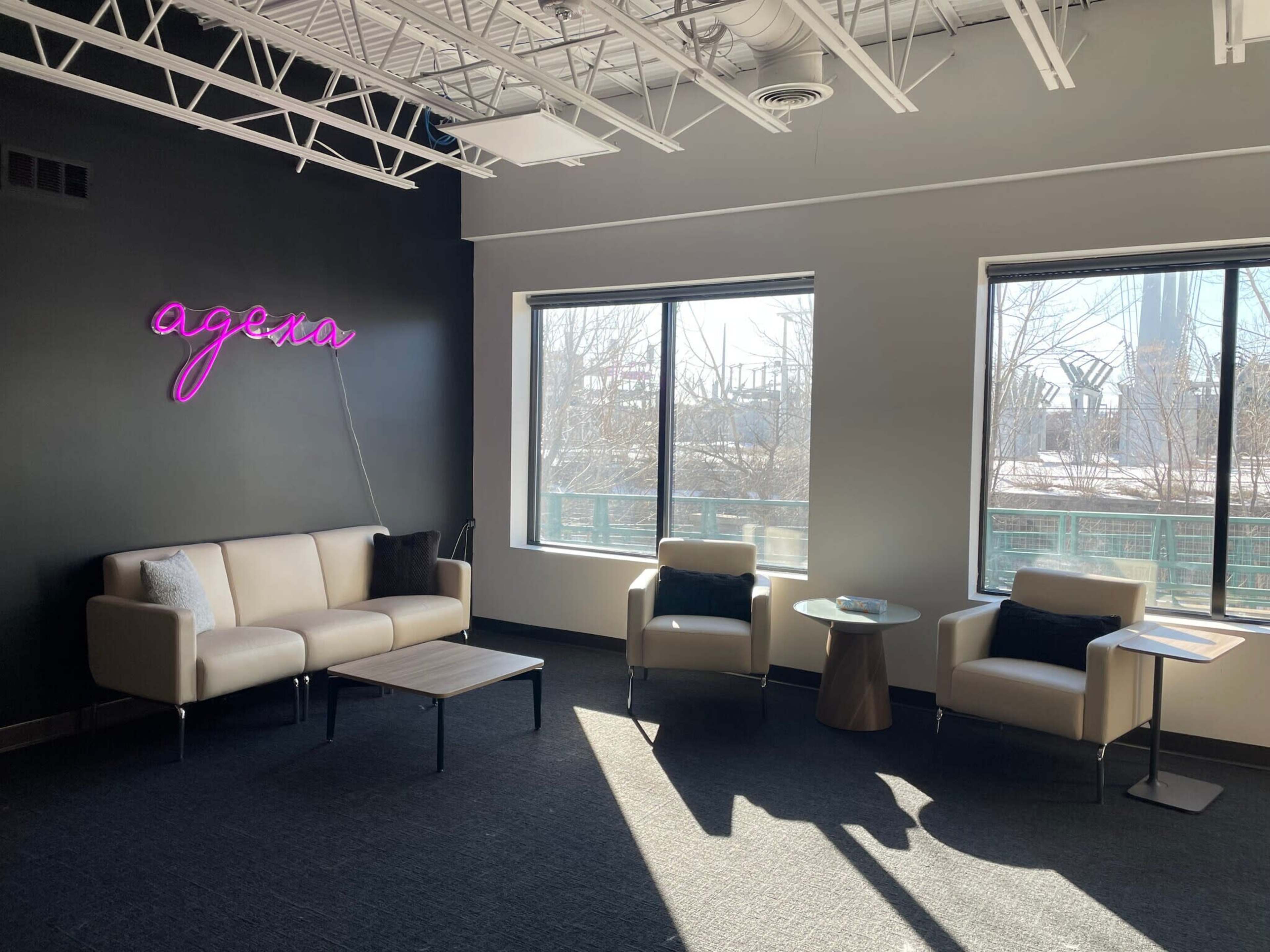The image shows a modern lounge area with a beige sofa, two white armchairs, a small table, and a wall featuring a neon "agenza" sign next to large windows.