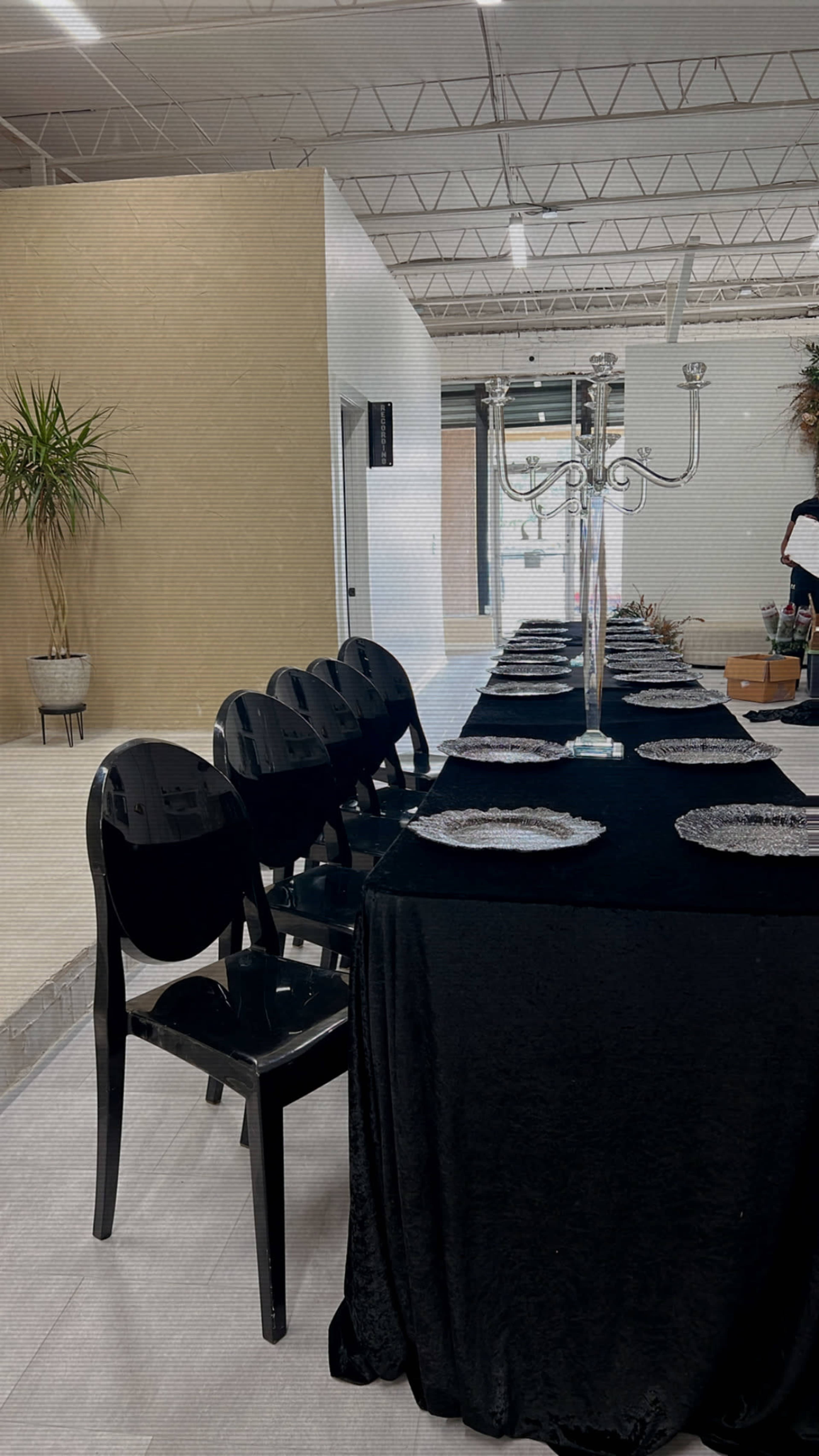 A long black table is set with plates and a candelabra, surrounded by black chairs in a spacious, minimally decorated room.