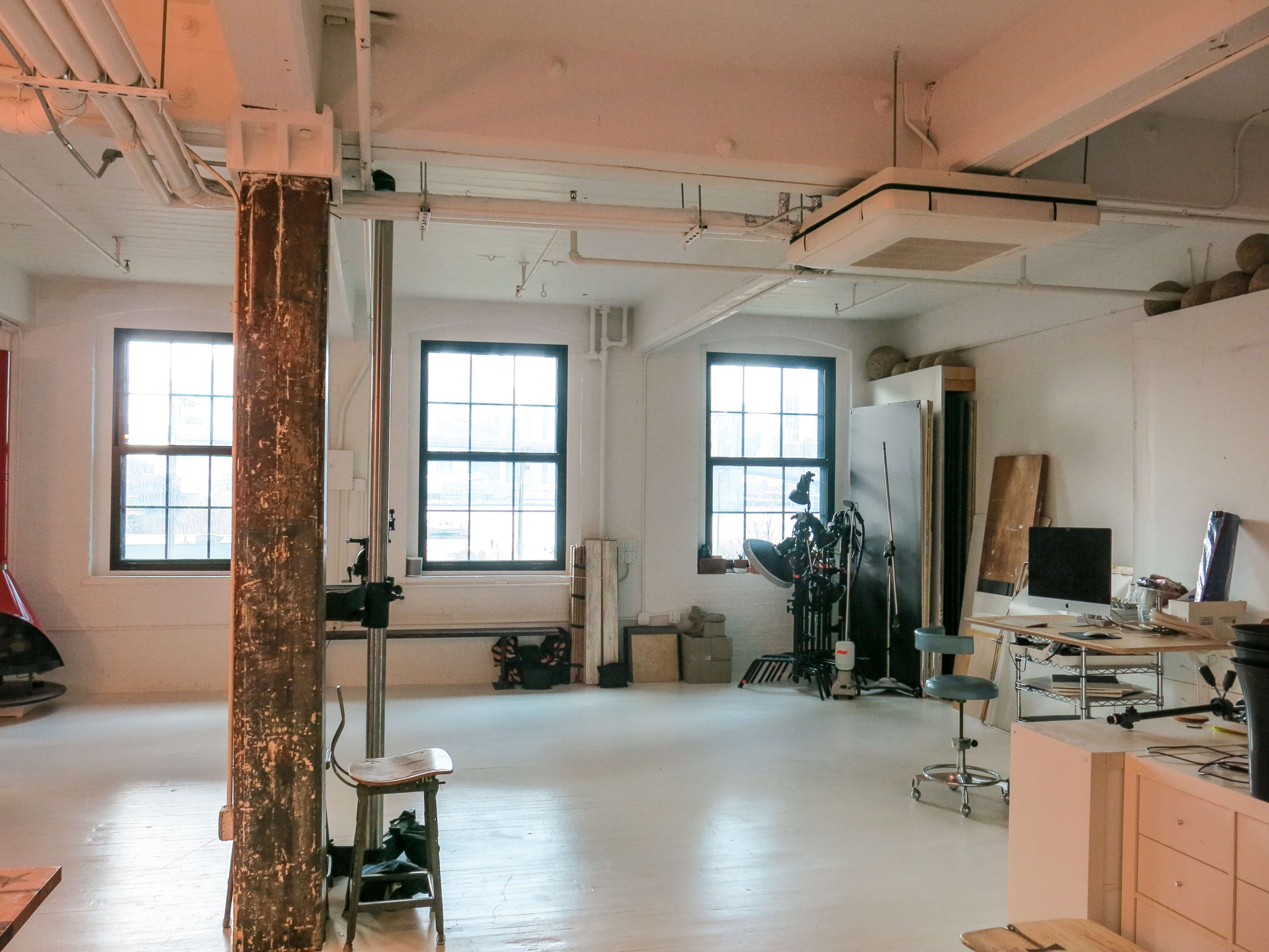 A bright, open studio space features large windows, a wooden pillar, various work surfaces, and art tools arranged against the walls.