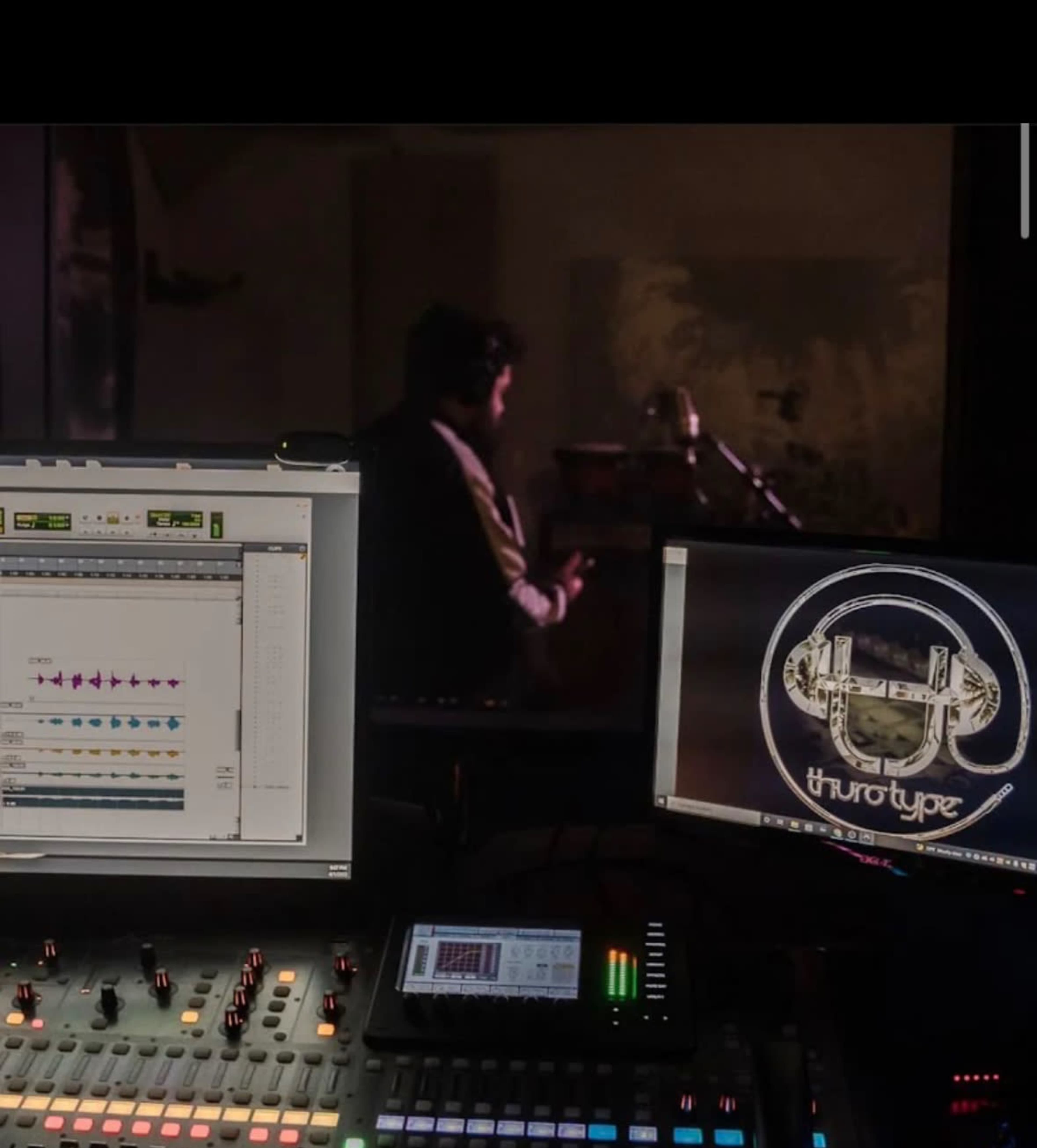 A recording studio setup features two computer monitors displaying audio editing software and a logo, with a musician partially visible in the background.