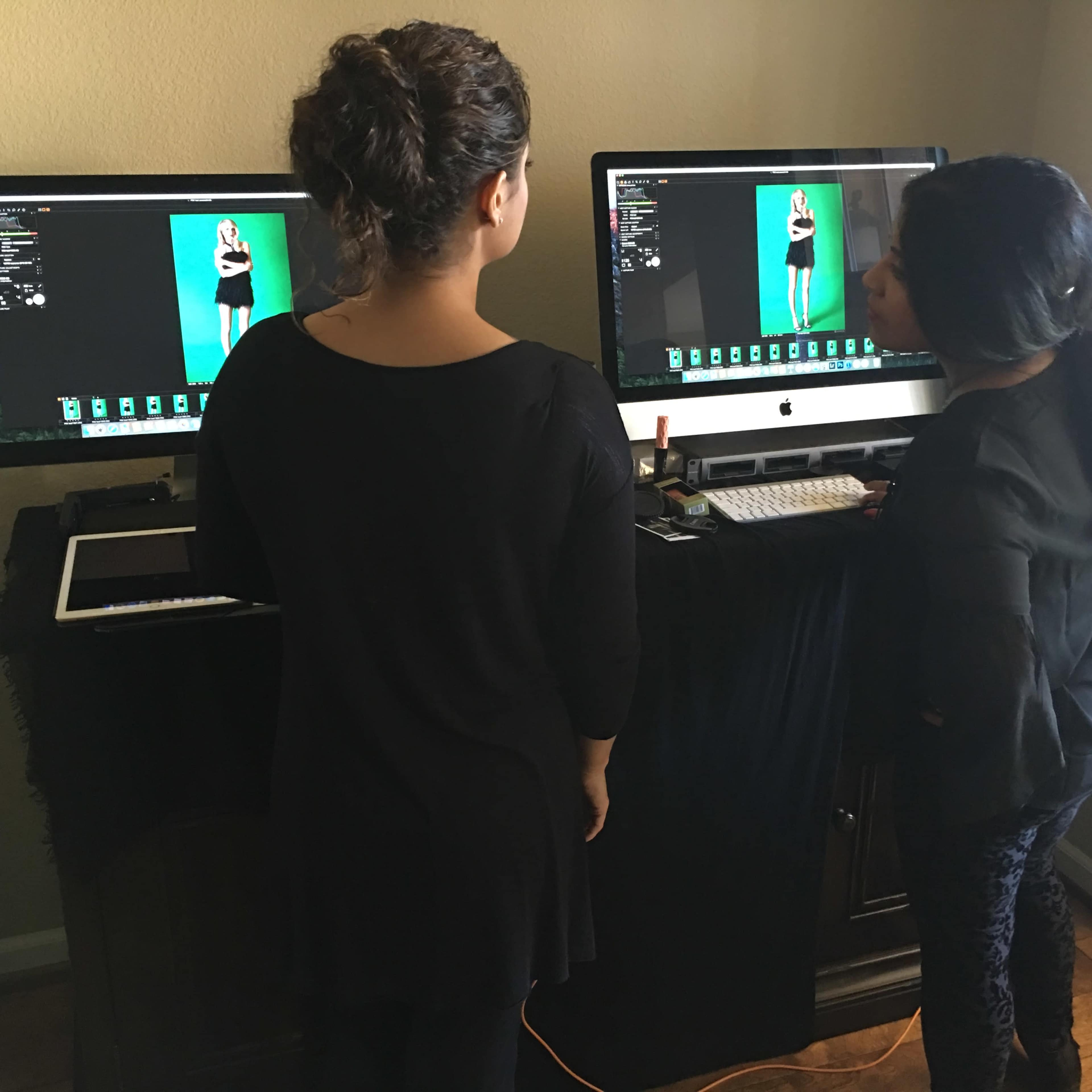 Two women are reviewing photos on computer screens with a green backdrop in a workspace.