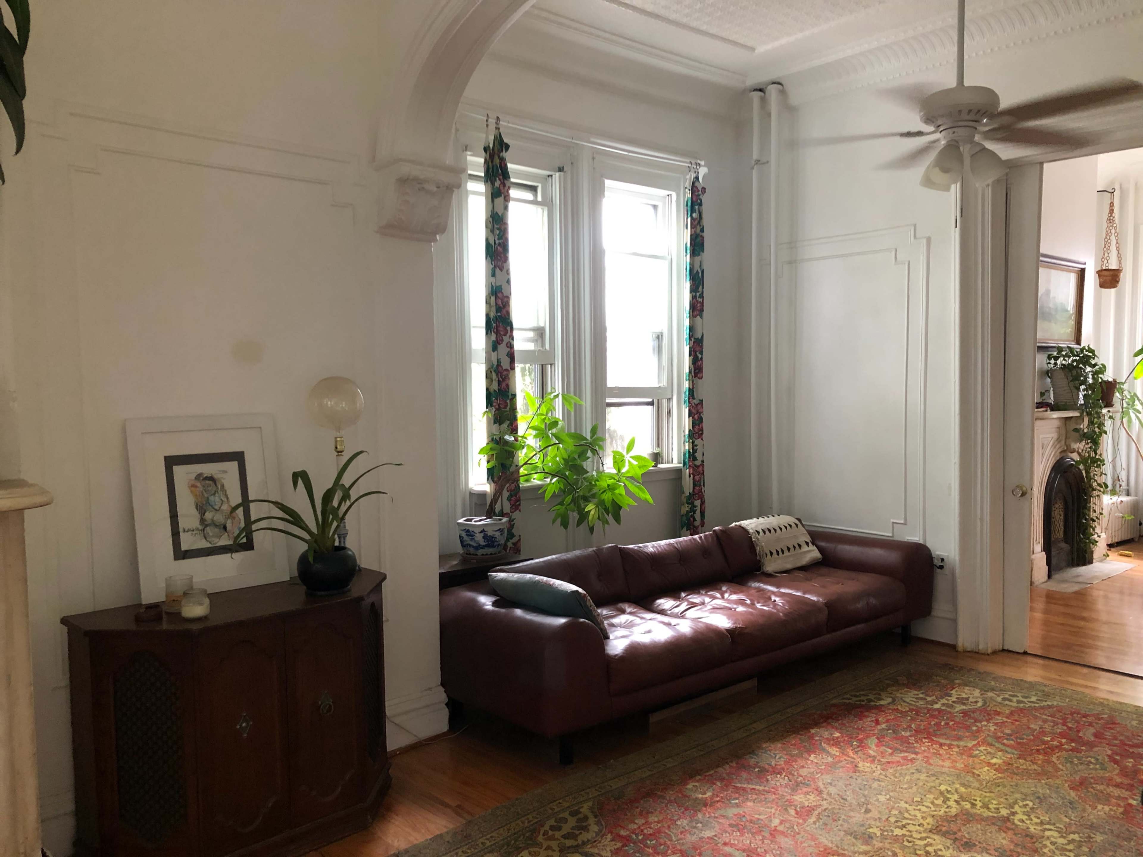 A living room featuring a brown leather sofa, a decorative rug, and large windows with plant accents.