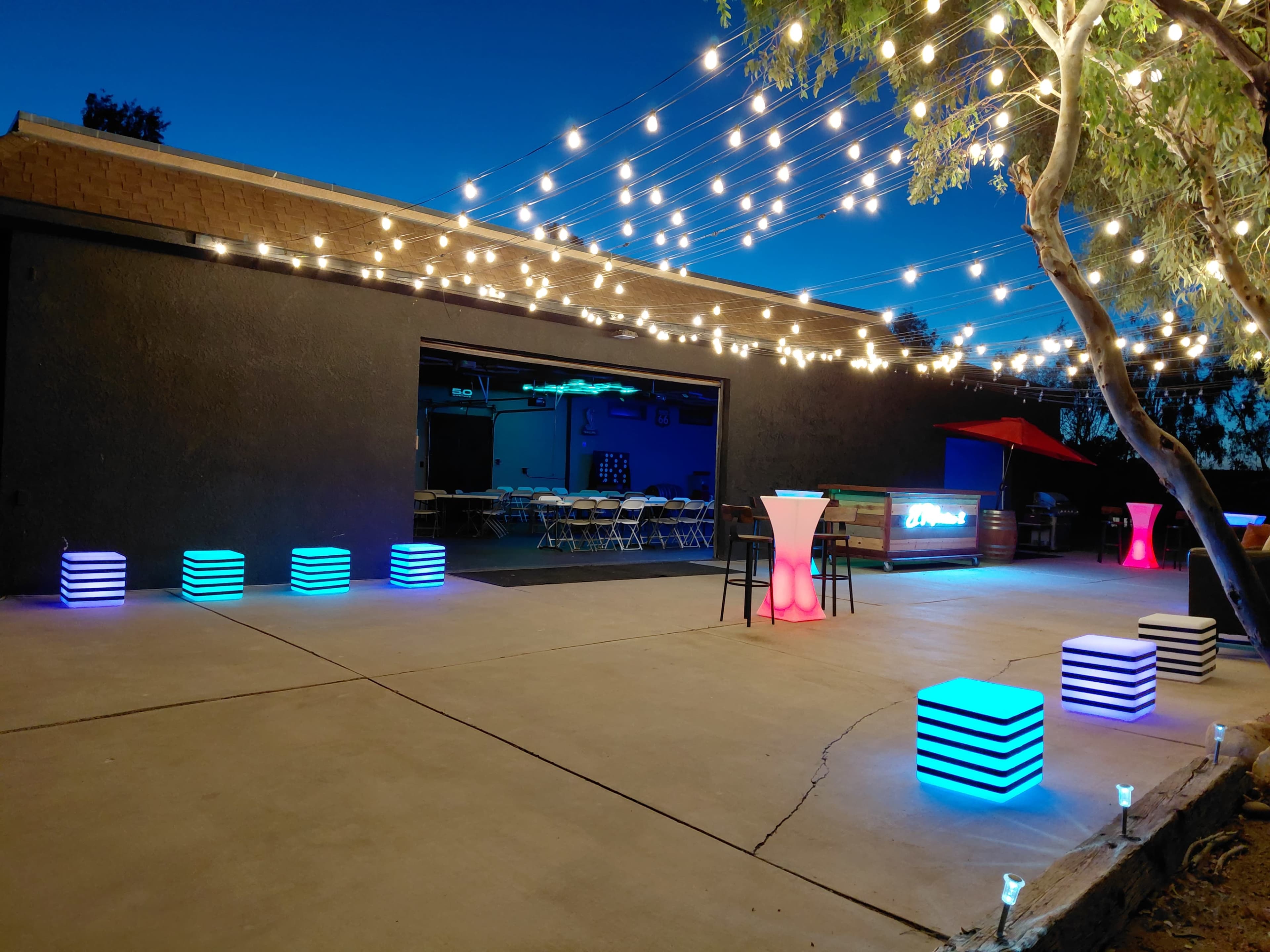 The image shows an outdoor event space featuring illuminated seating cubes, string lights overhead, and a bar area next to a garage opening.