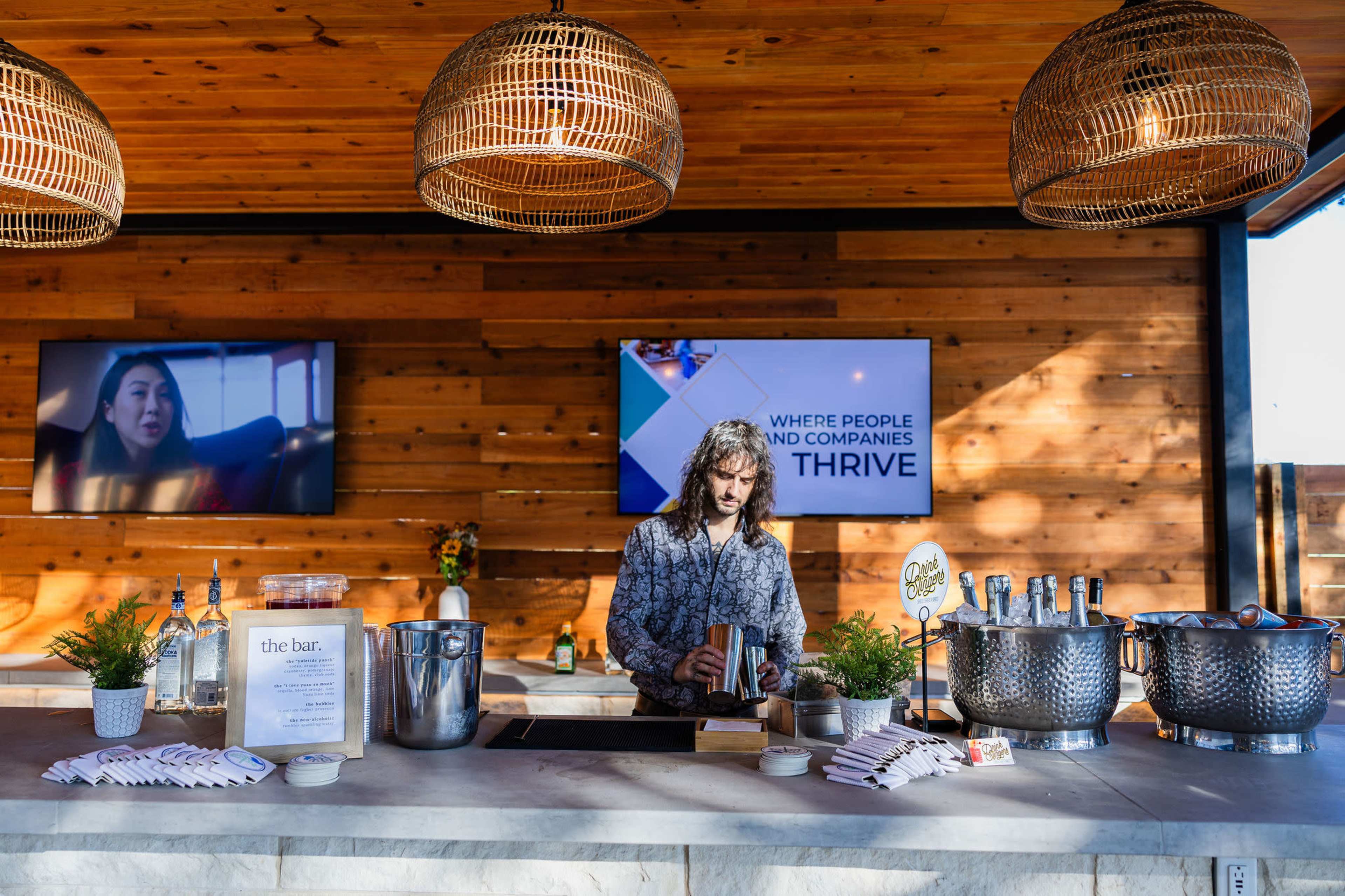 A bartender prepares drinks at a stylish counter beneath hanging light fixtures, with screens displaying messages in the background.