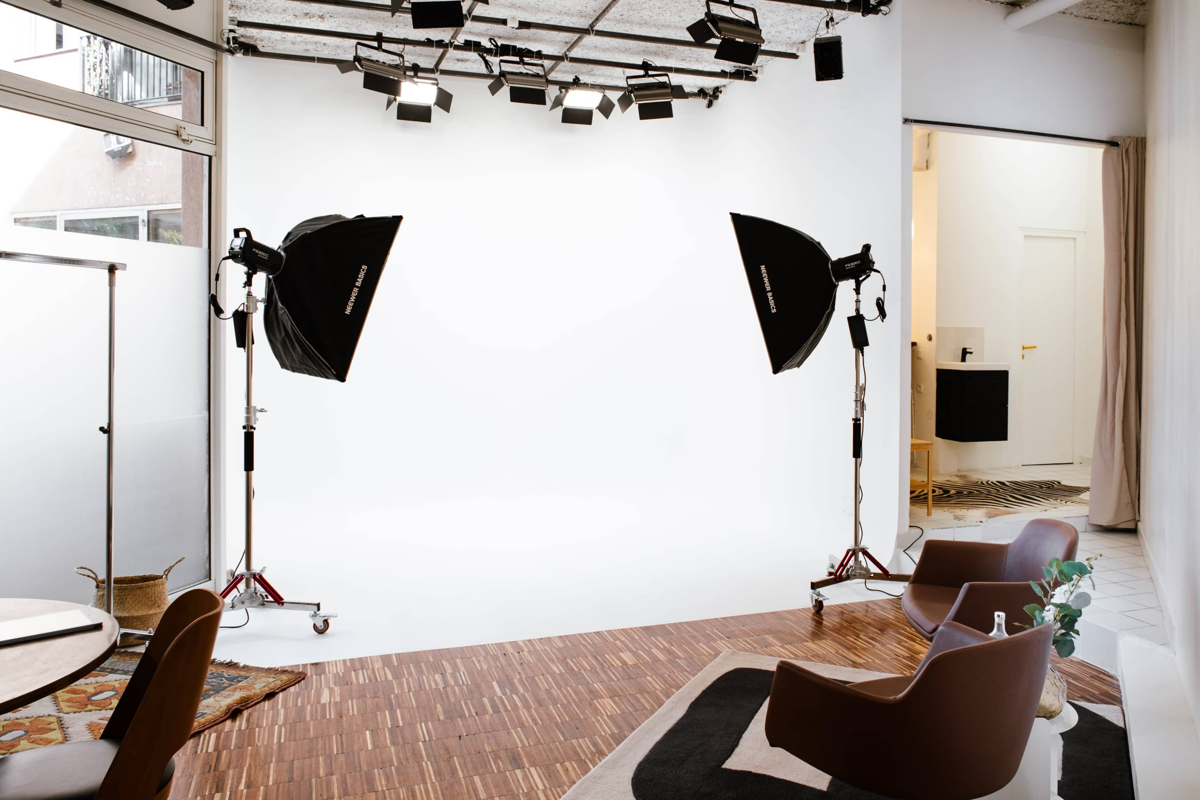 The image shows a photography studio with two large softbox lights, a clean white backdrop, and modern seating arrangements.