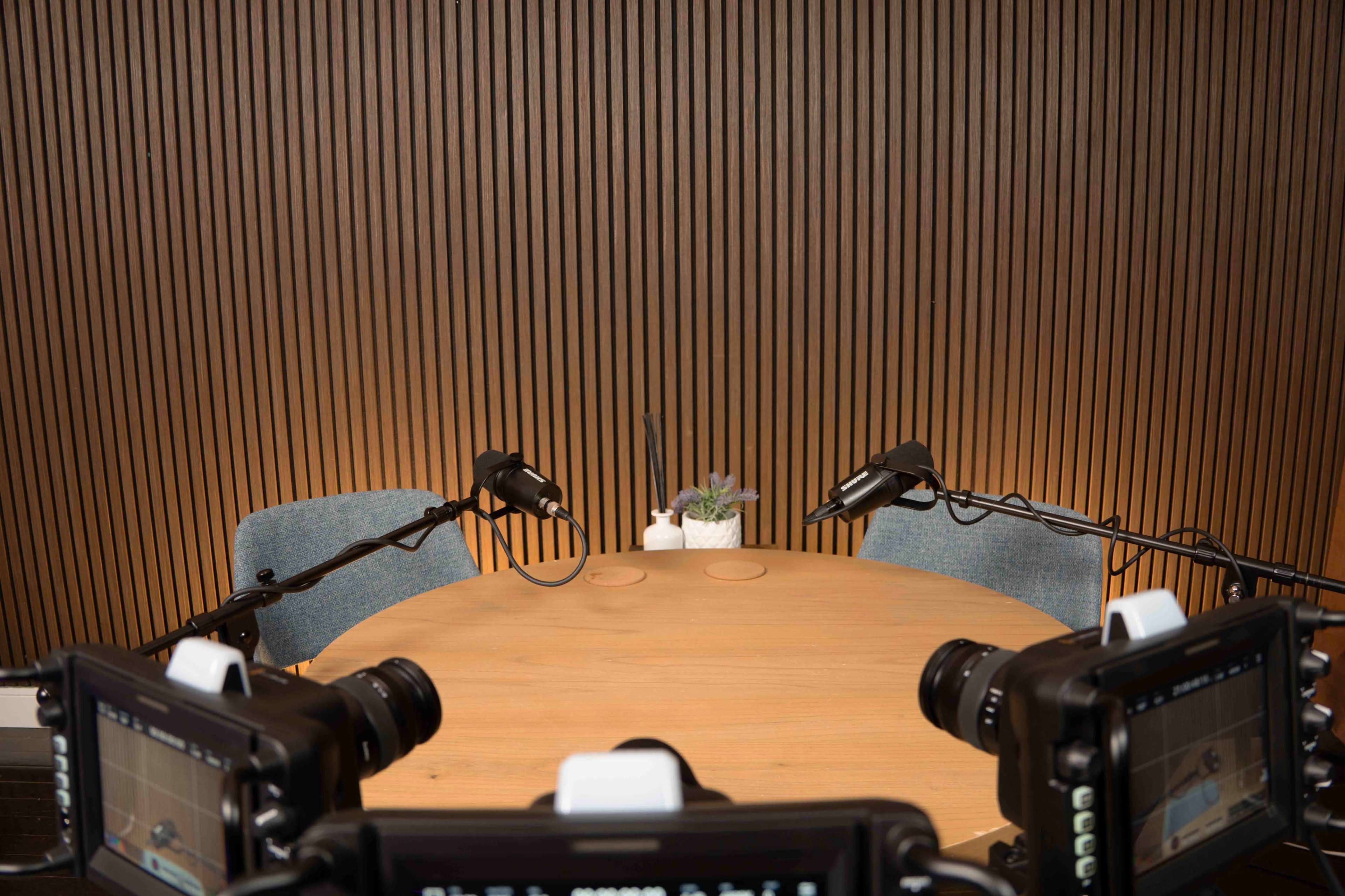 A wooden table set with two chairs is surrounded by cameras and microphones, with a small plant at the center.