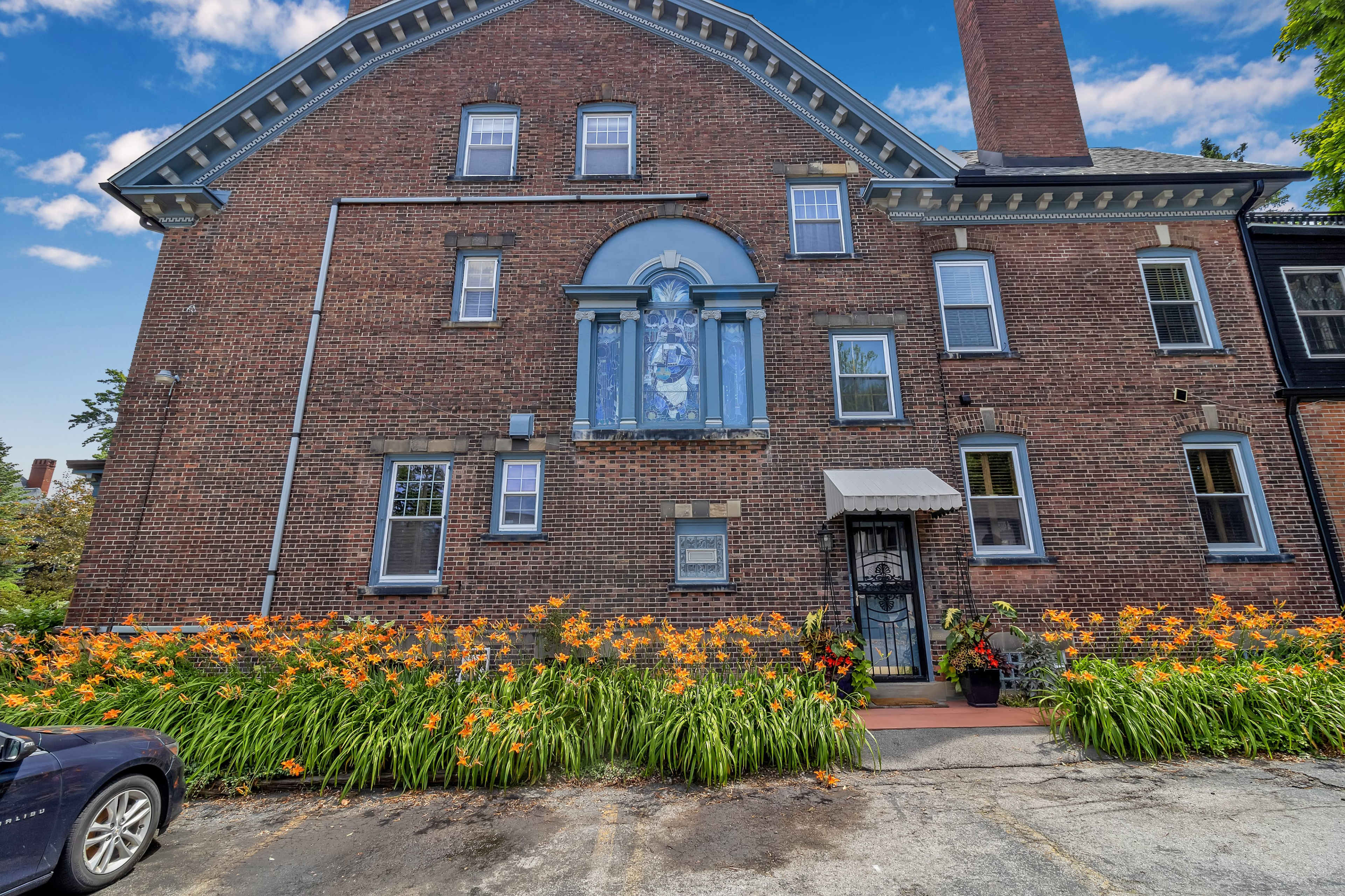 A brick building features a large blue window and is surrounded by a bed of orange daylilies, with a vehicle parked nearby.