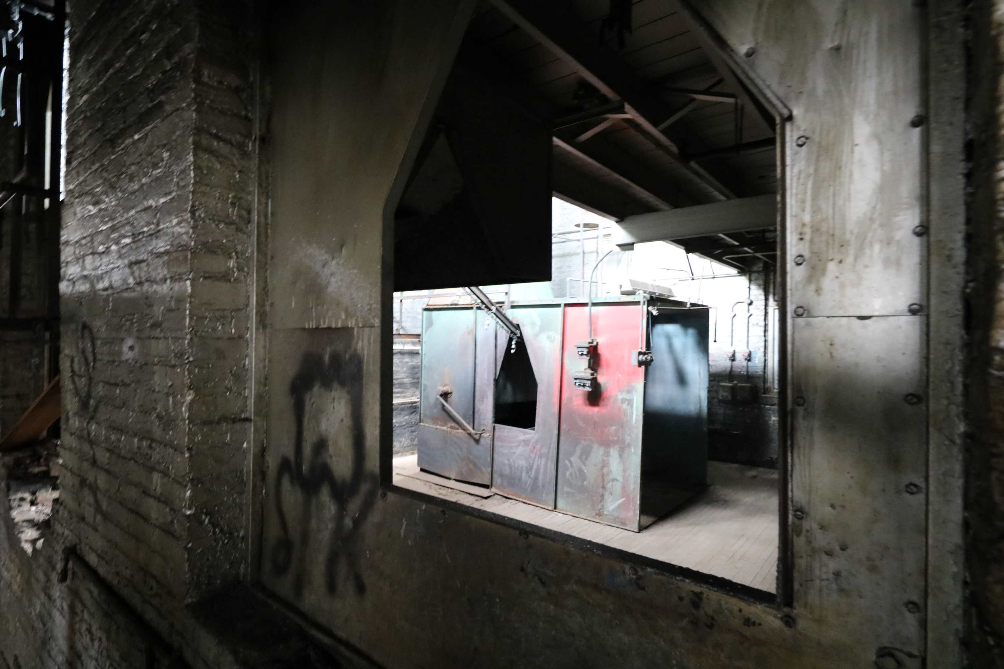 The image shows a view through a broken window into an abandoned industrial space featuring two metallic containers against a wall.