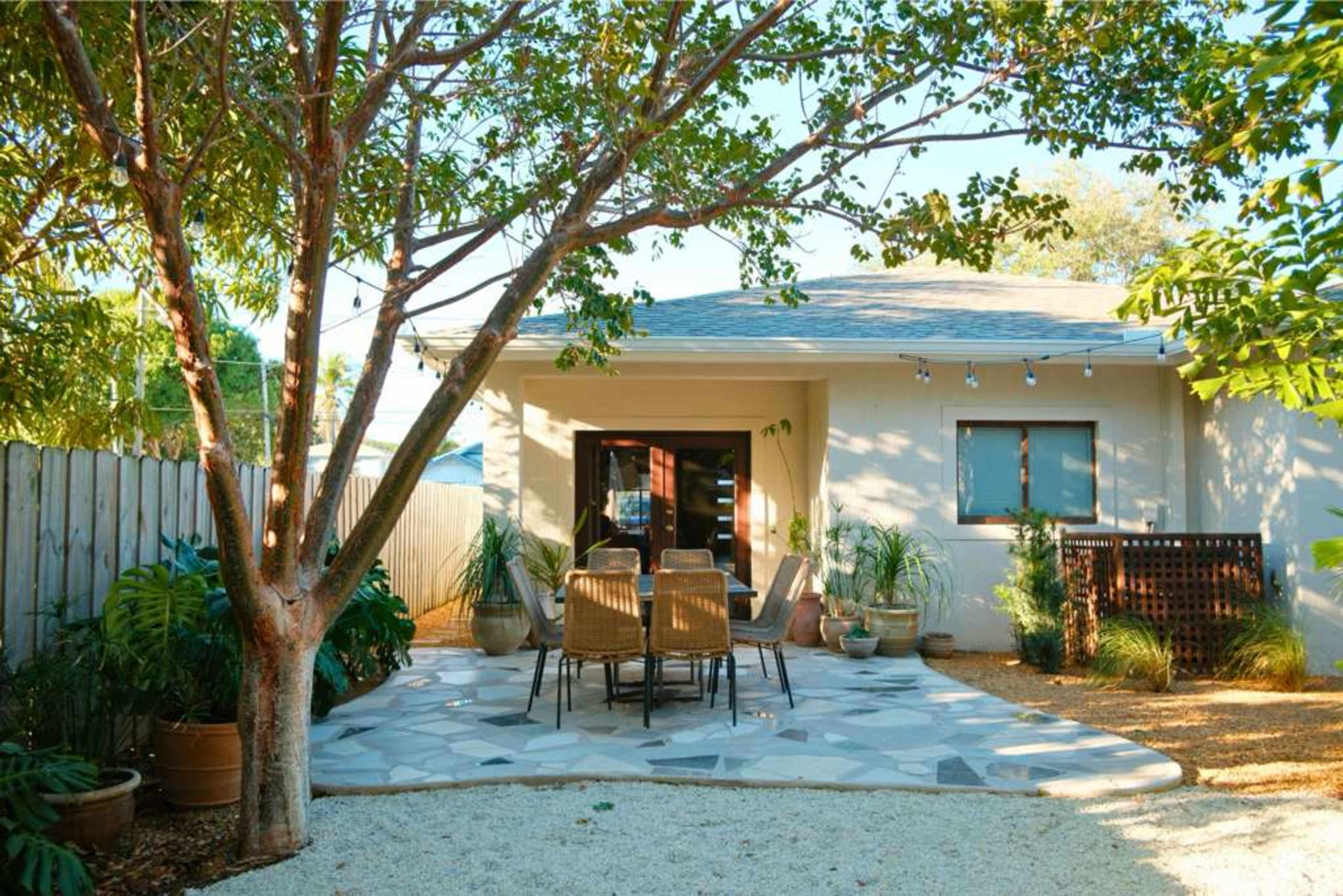 The image shows a small patio area with a stone floor, a table and chairs, surrounded by greenery and a tree.