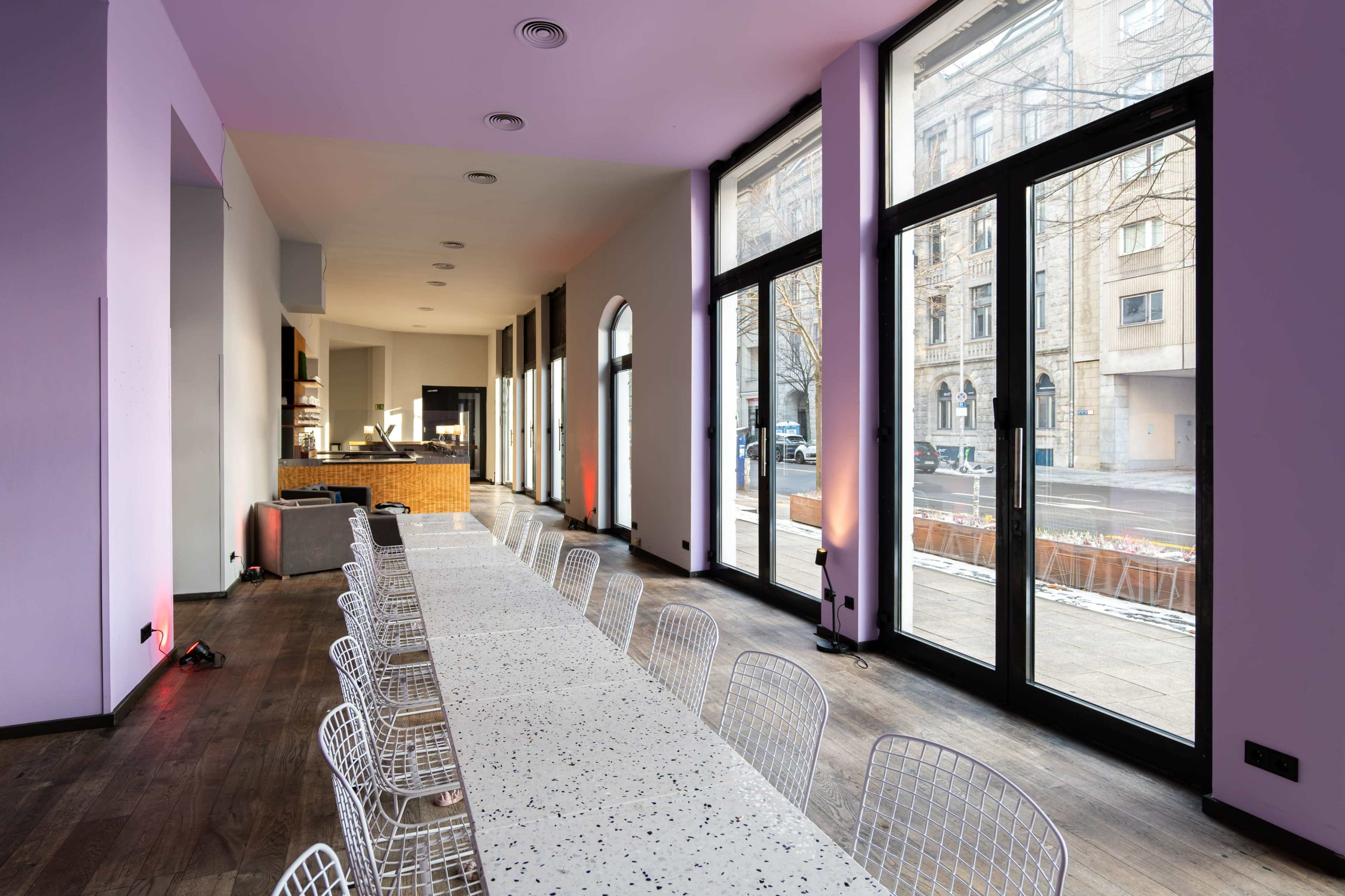 A long table with white chairs is set in a brightly lit room featuring large windows facing an urban street.