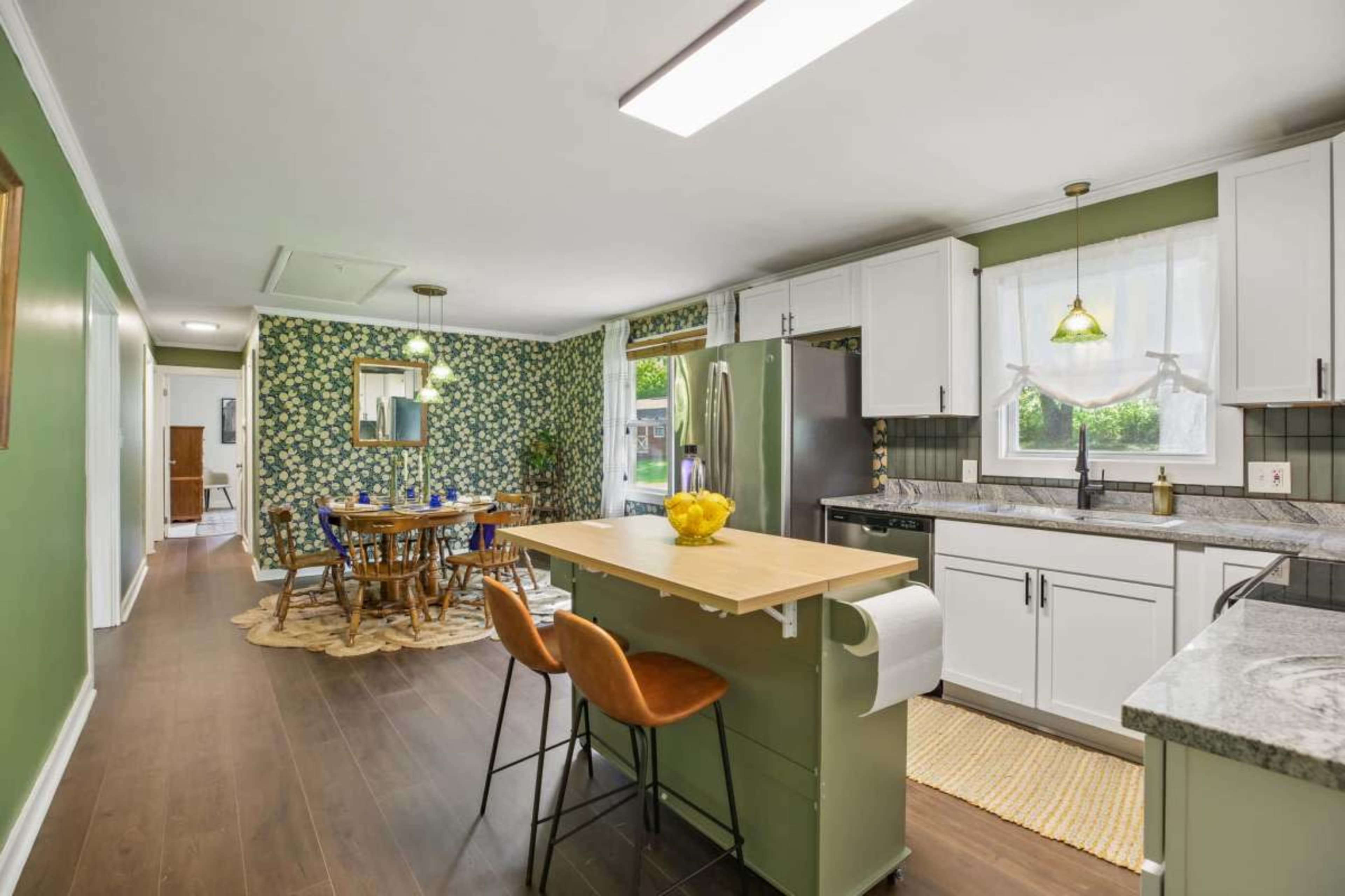 The image shows a modern kitchen with white cabinets, a large island with bar stools, and a dining area featuring a round table set for a meal against floral wallpaper.