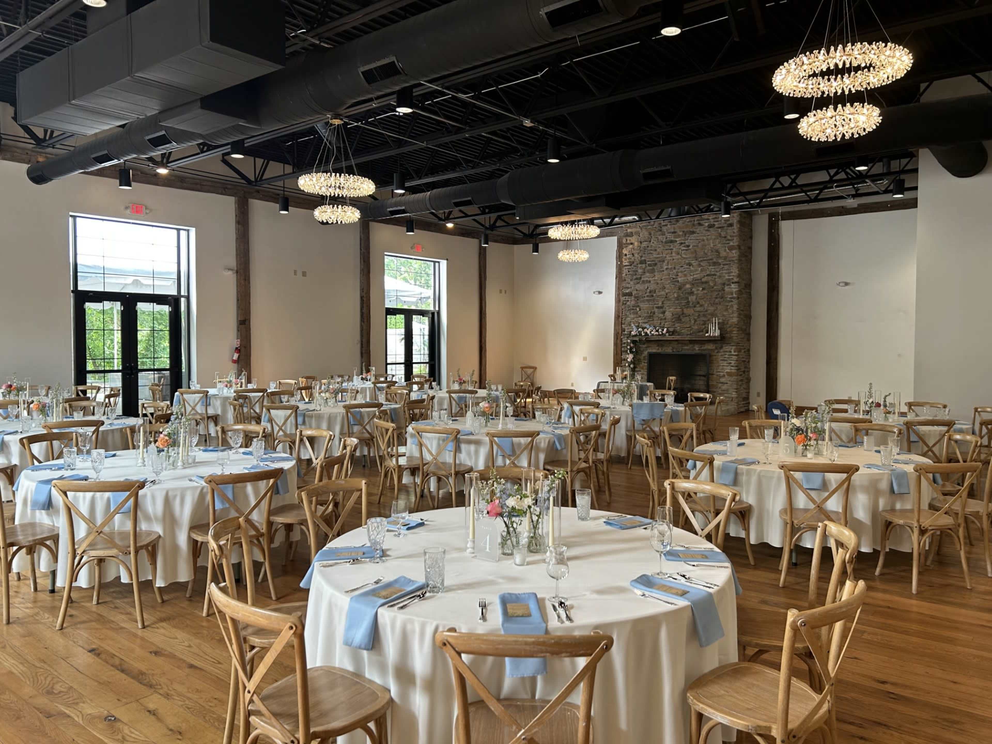 The image features a dining area set up for an event, with multiple round tables covered in white tablecloths, wooden chairs, and decorative centerpieces, all under a high ceiling with modern chandeliers.