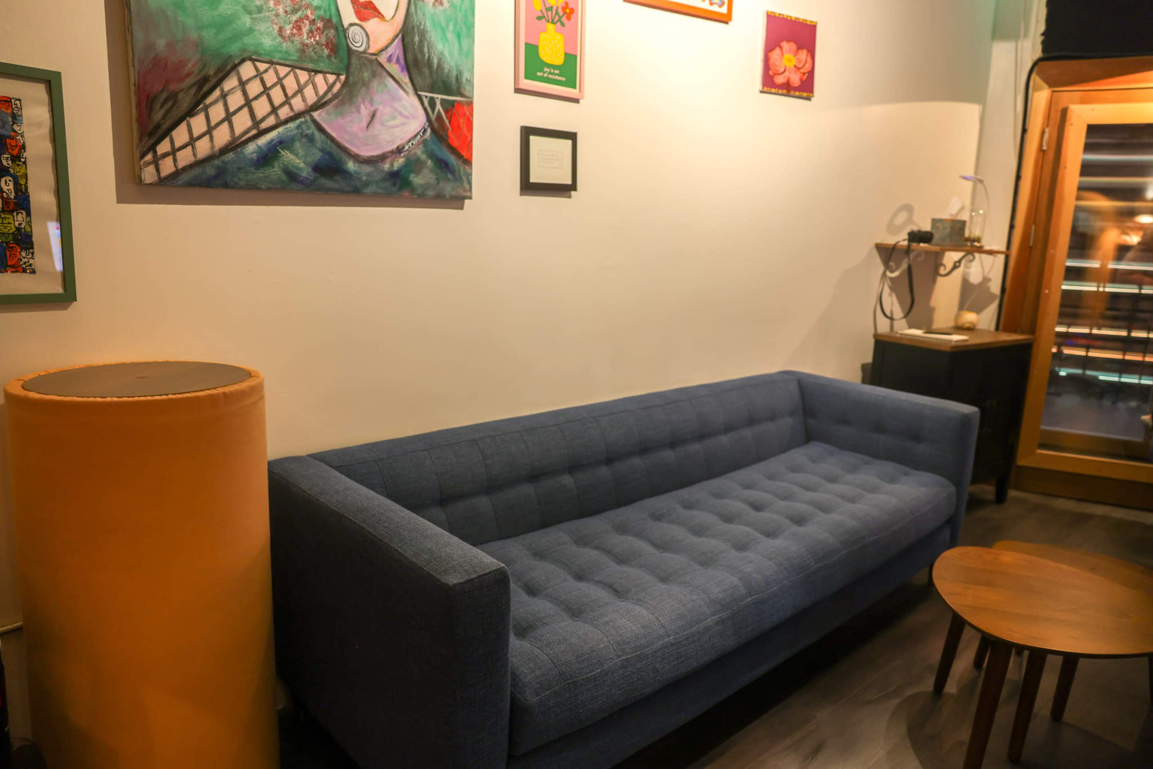 The image shows a blue tufted sofa positioned against a white wall adorned with colorful artwork, accompanied by a small round wooden table and a yellow cylindrical lamp.