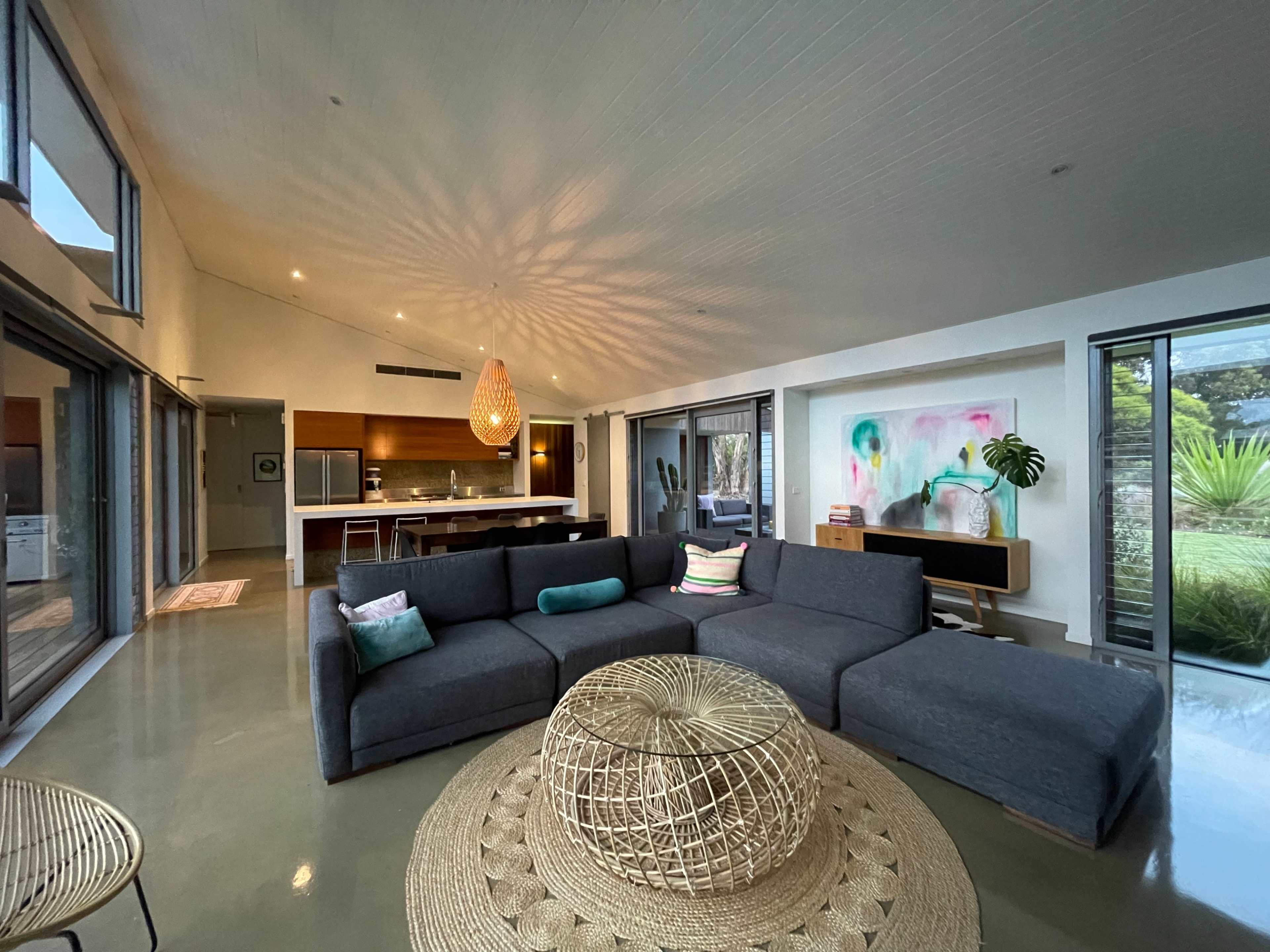 The image shows a modern living room with a large sectional sofa, a round coffee table, and a kitchen visible in the background, featuring large windows that provide natural light.