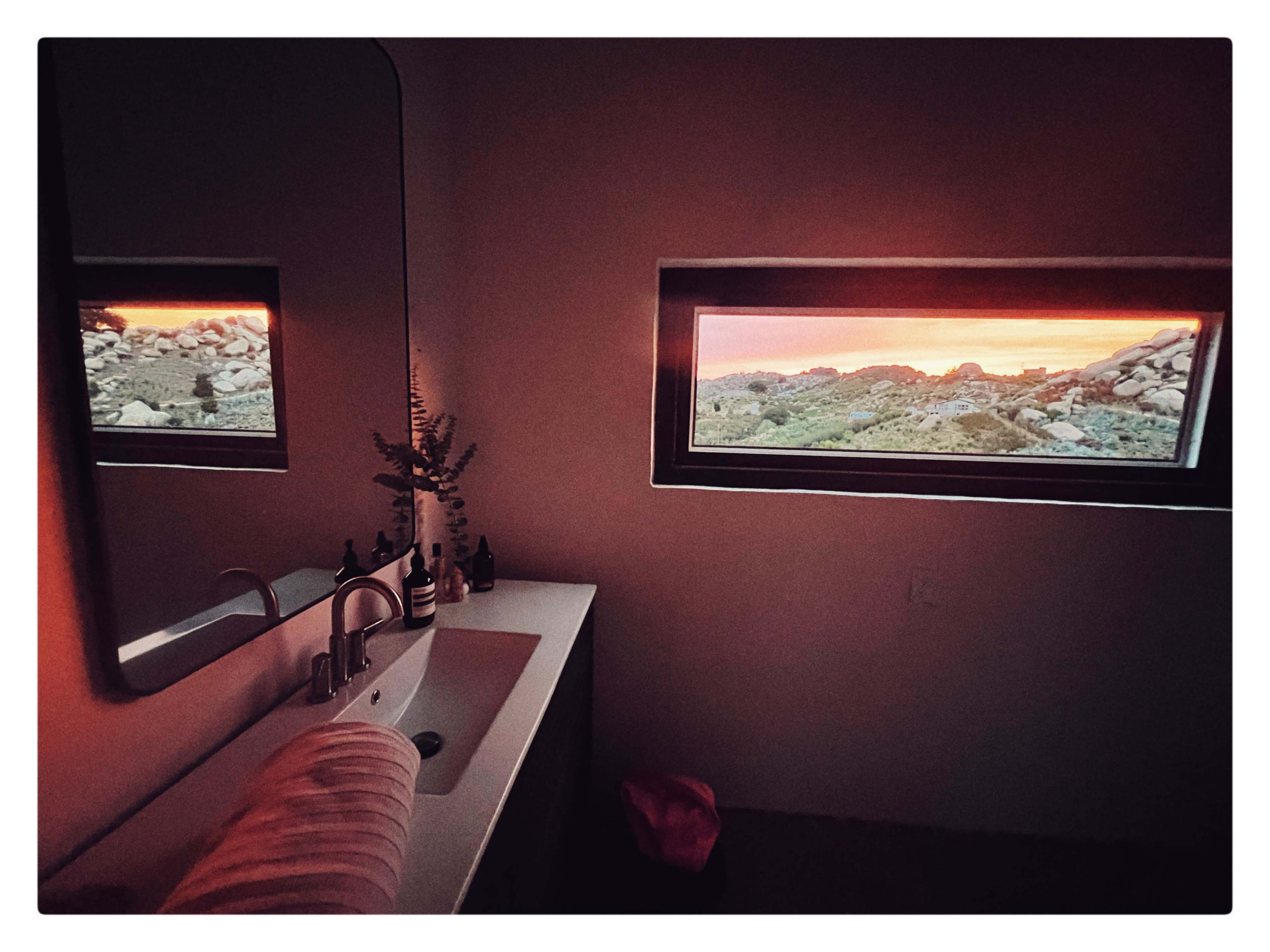 A modern bathroom features a sink and mirror, with a large window showcasing a vibrant sunset over rocky terrain.