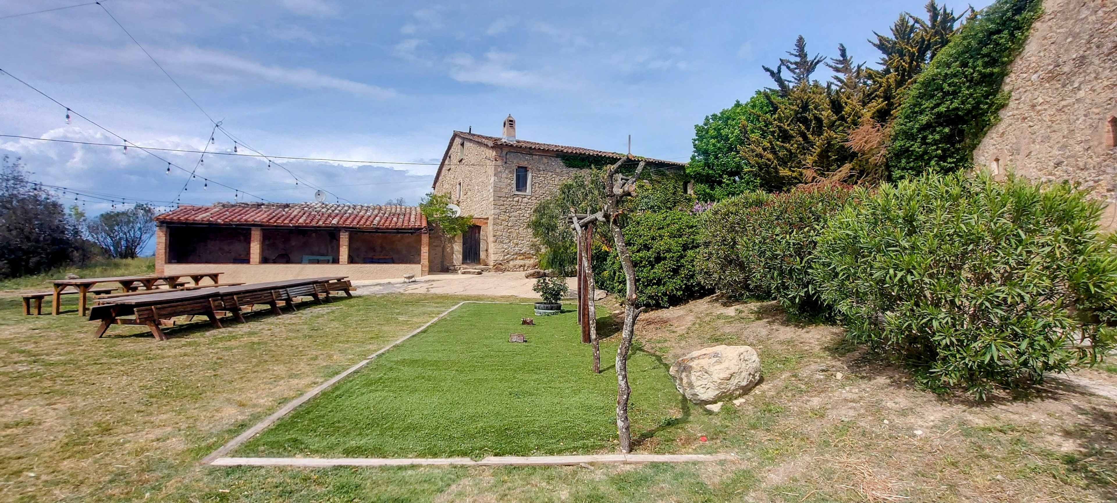The image shows a rustic farmhouse with a stone exterior, surrounded by greenery and a small lawn area with wooden benches and a gravel patio.