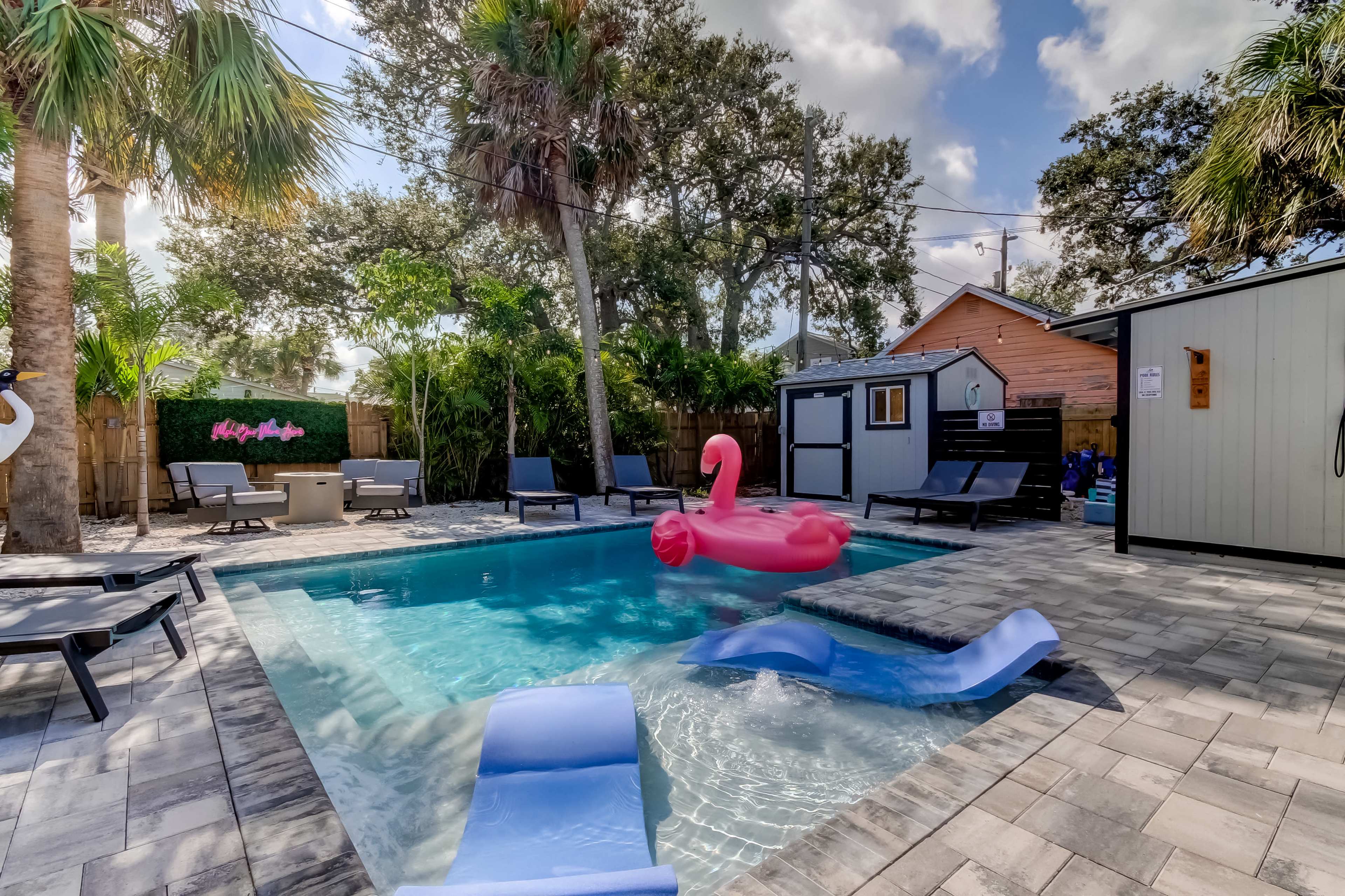 A backyard pool area featuring a pink flamingo float, sun loungers, and tropical plants surrounding the pool.