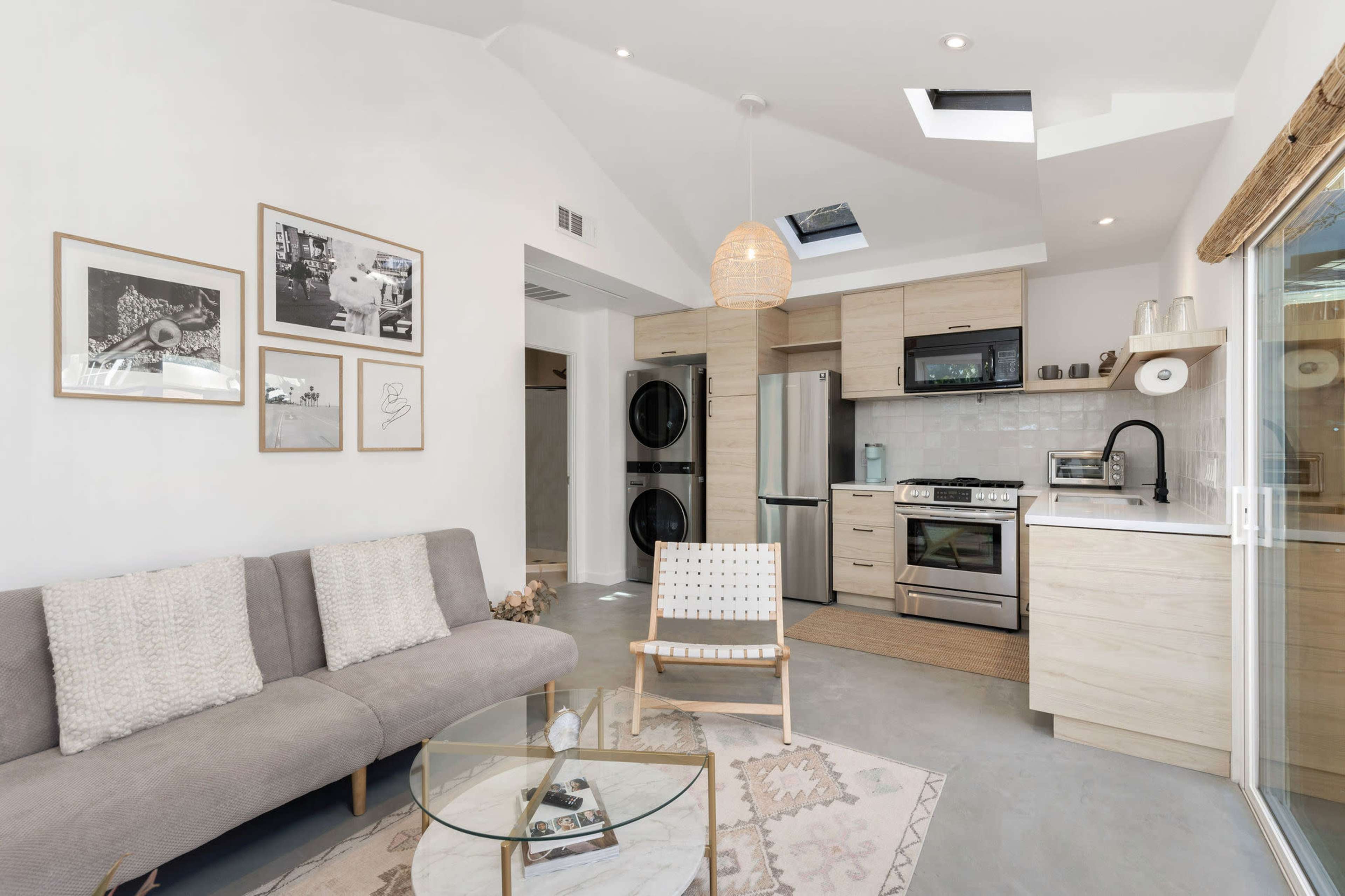 A modern living space featuring a light-colored sofa, a round glass coffee table, and a kitchen area with stainless steel appliances.