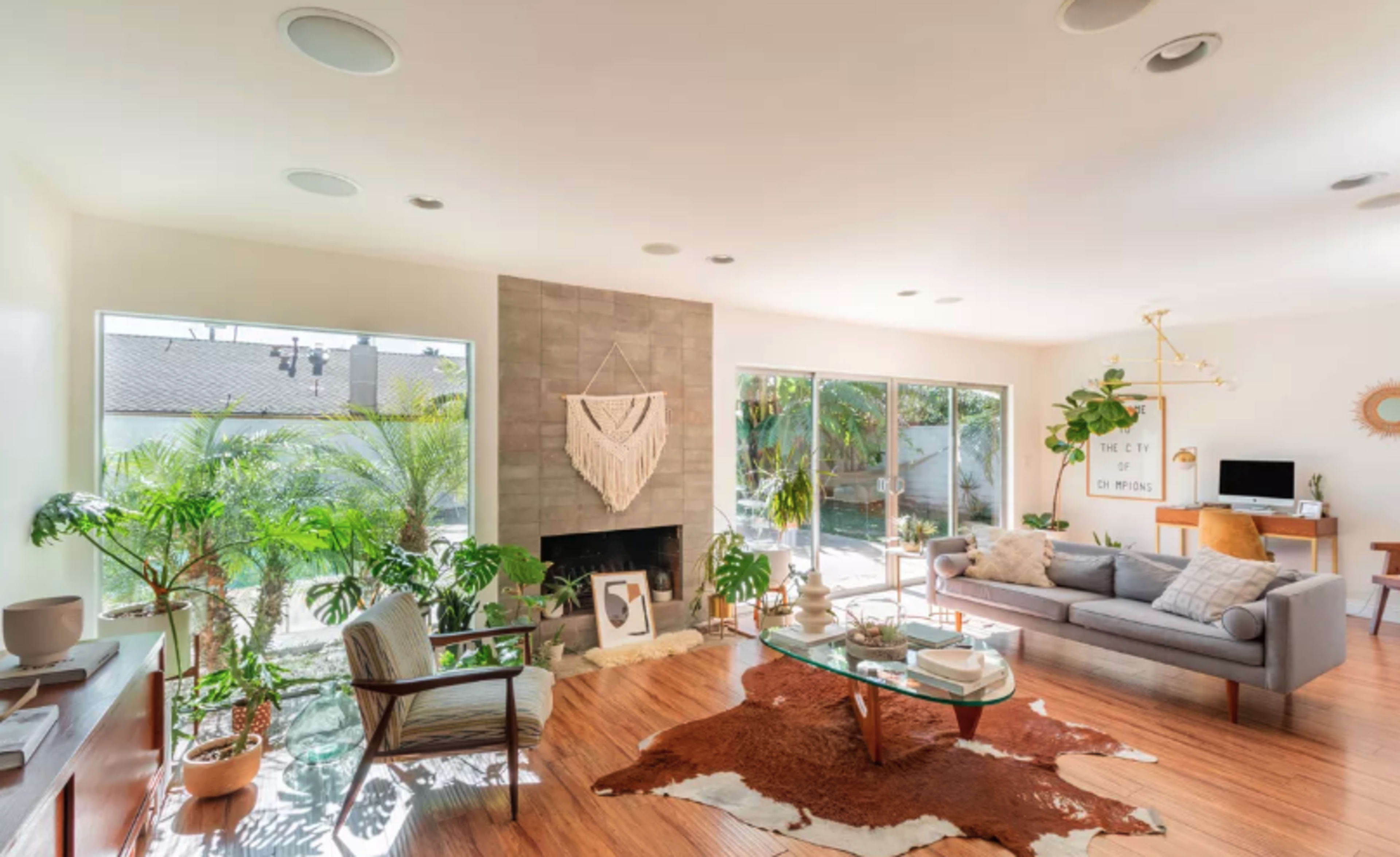 The living room features a grey sectional sofa, a round glass coffee table, and large windows that overlook a patio with greenery.